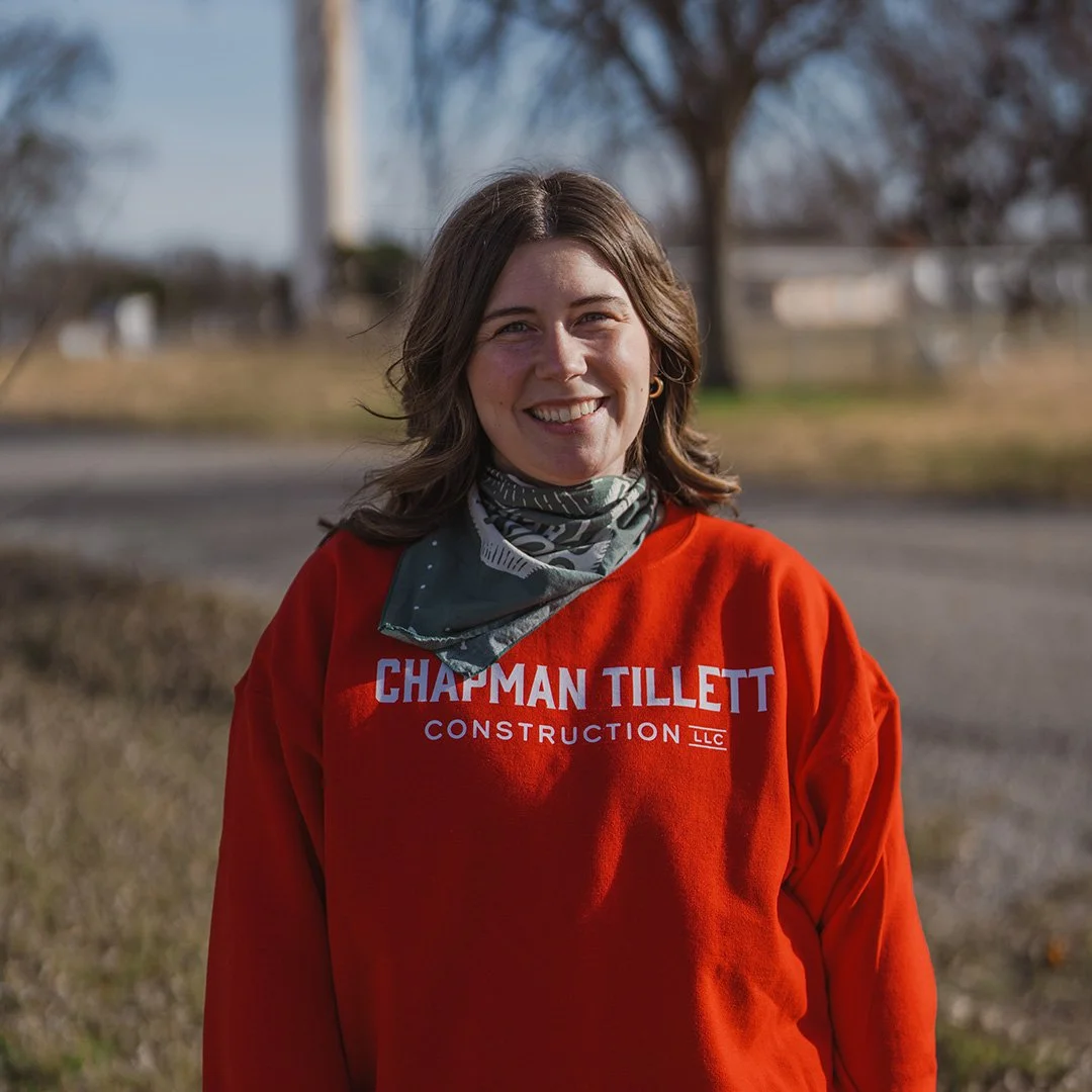 A woman with shoulder-length wavy brown hair smiling outdoors, wearing a tan crochet top and a dark green scarf with white patterns, in a natural setting with trees in the background.