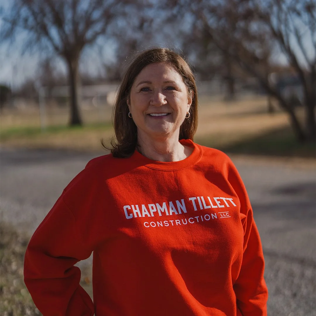 A woman with shoulder-length brown hair, wearing a bright pink satin blouse, sitting at a wooden table and smiling at the camera.