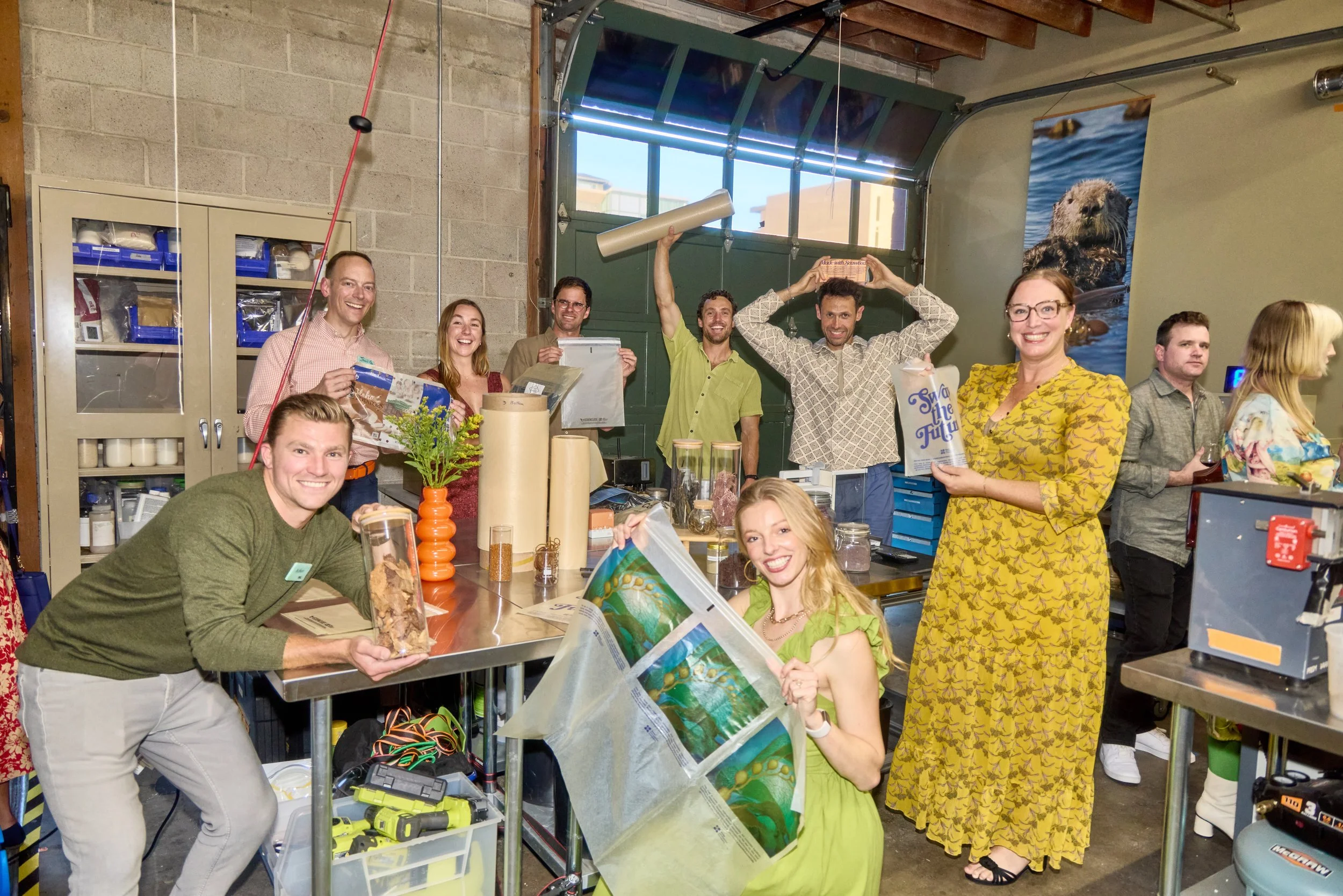 A group wearing Unwaste reusable name tags poses with Sway's compostable seaweed-based packaging samples at a joint sustainability event, highlighting a partnership between two material innovators.
