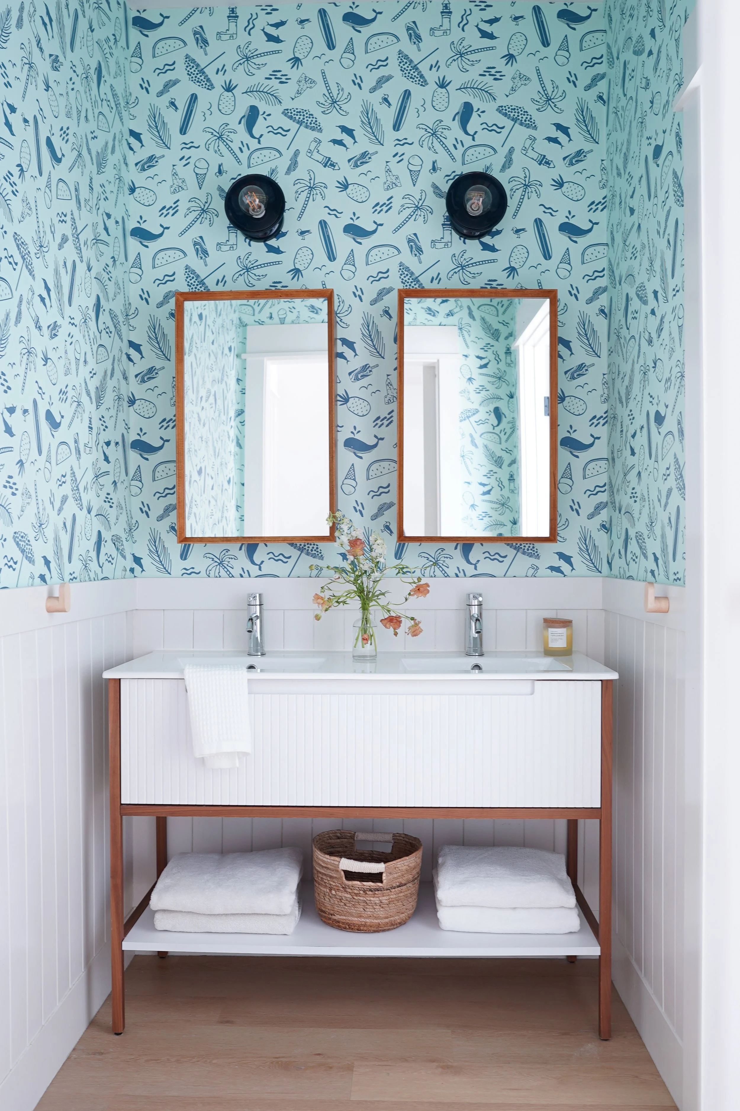 A bathroom with a blue wallpaper featuring water-themed illustrations like whales, dolphins, palm trees, and ice cream. Two mirrors with wooden frames are mounted above a white sink with two faucets. Below the sink, a wooden frame structure holds nea