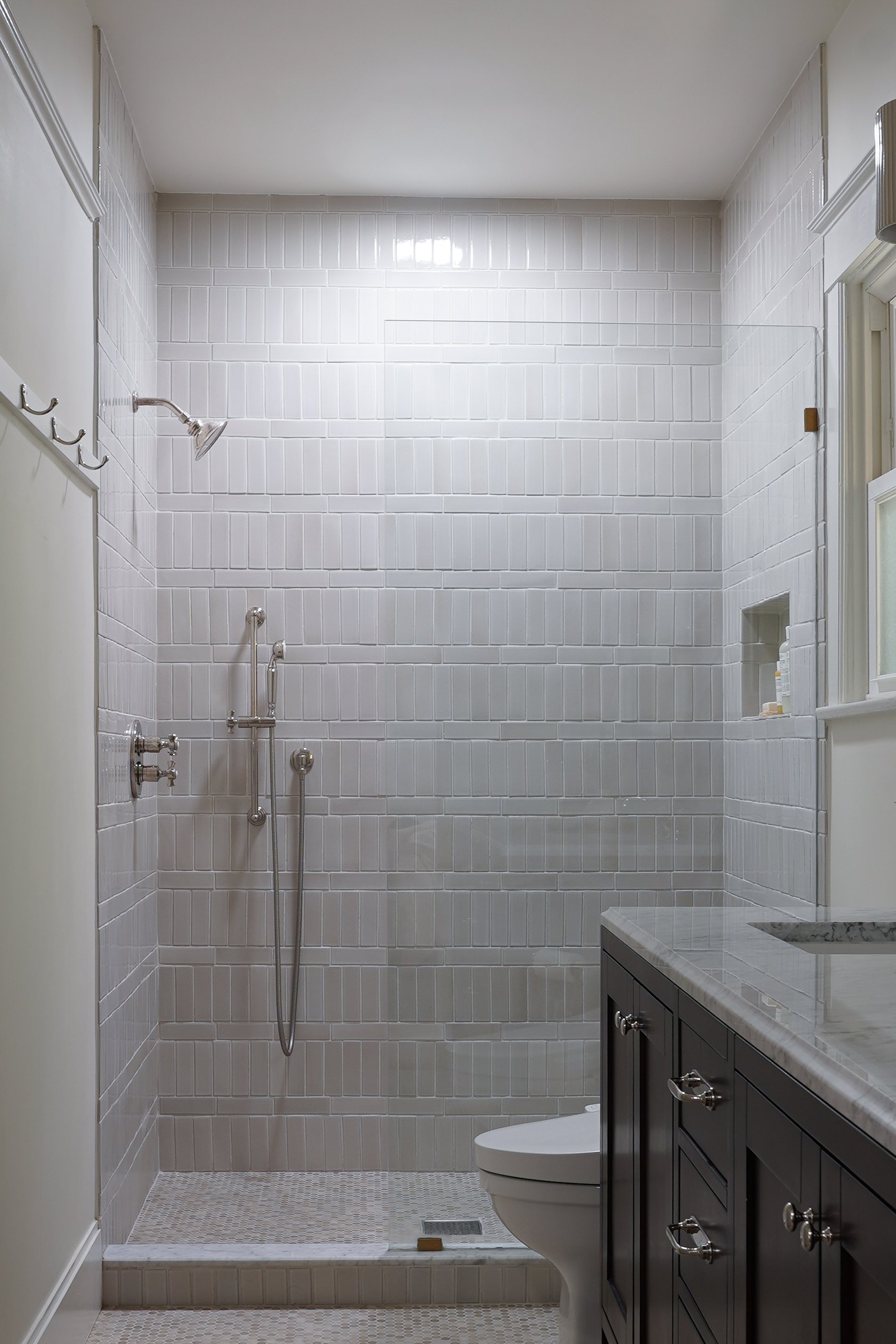 Bathroom with white tiled shower area, a dark wooden vanity with a marble countertop, and a toilet. The shower has a handheld showerhead and tiled walls.