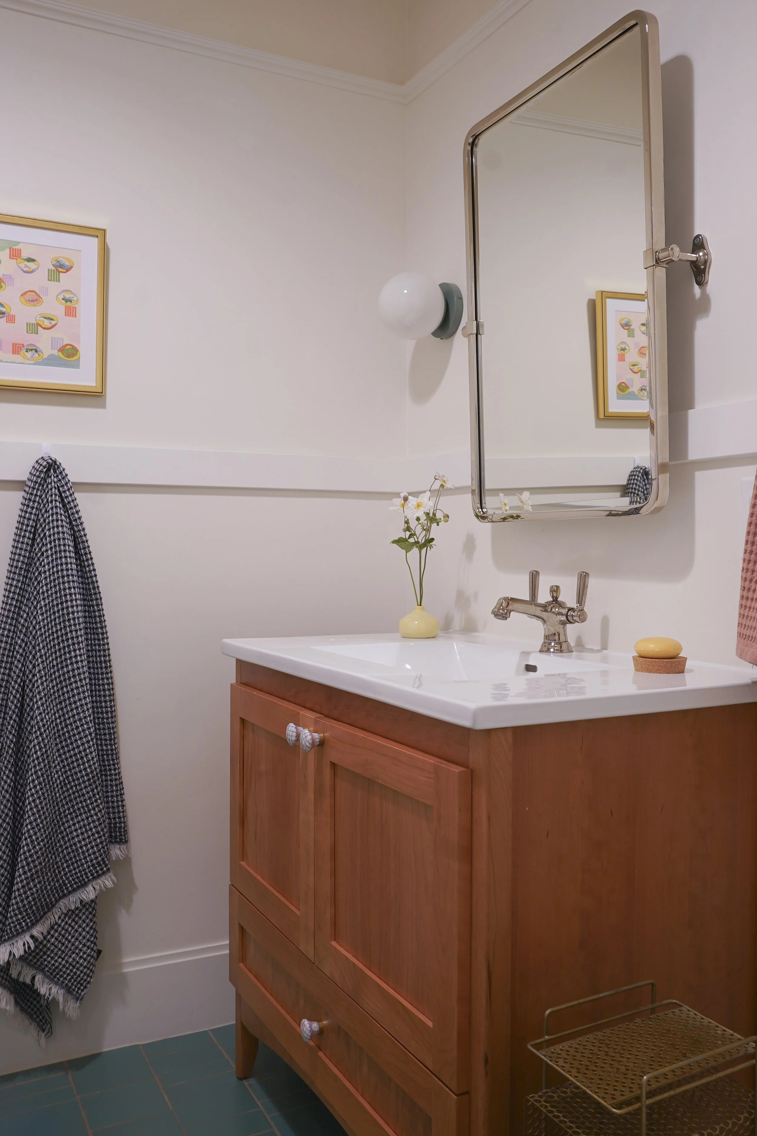 A bathroom with a wooden vanity, white countertop, and a faucet. Above the sink is a round mirror with a metal frame. To the left, a checkered towel hangs on a hook. On the right, a wall-mounted light fixture. On the countertop, a yellow vase with wh