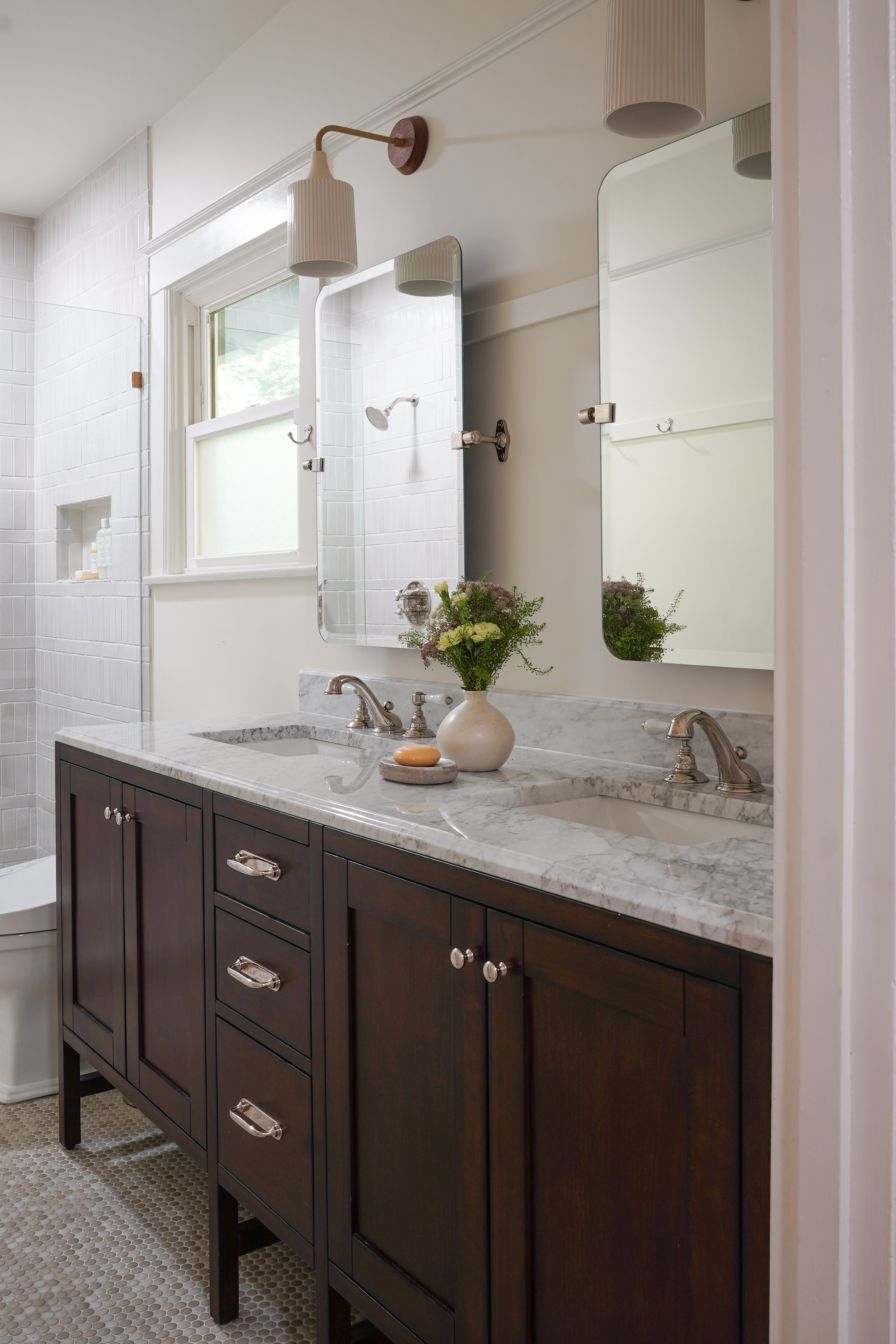 Bathroom with dark wooden vanity, white marble countertop, double sinks, and wall mirrors. Flowers in a vase, soap, and bathroom essentials near the window, with white tiled walls and a patterned floor.