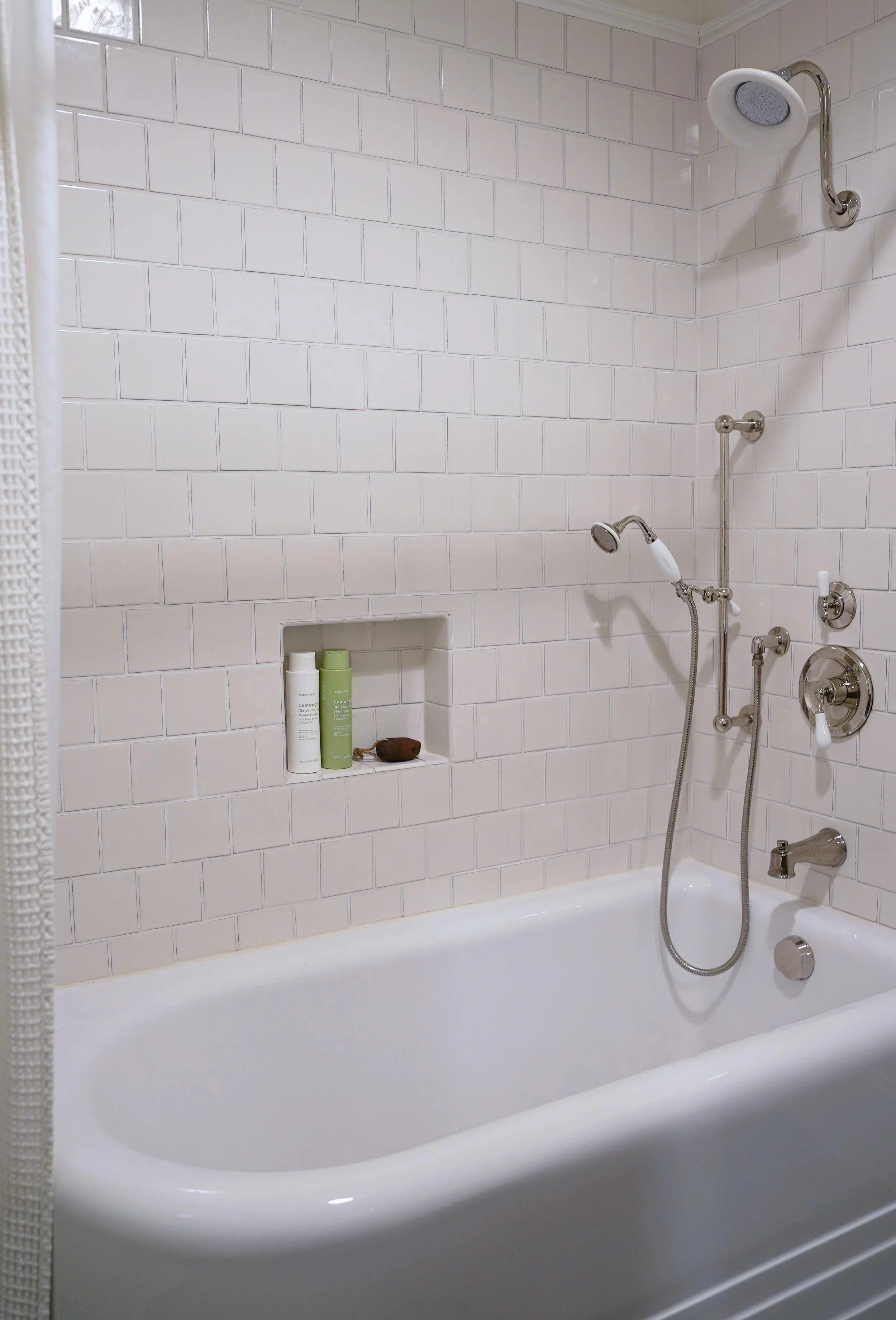 A white bathtub in a bathroom with white subway tile walls. There are two shower heads and multiple pipes, one on a vertical bar. A small built-in shelf in the wall holds bottles and a stone object.