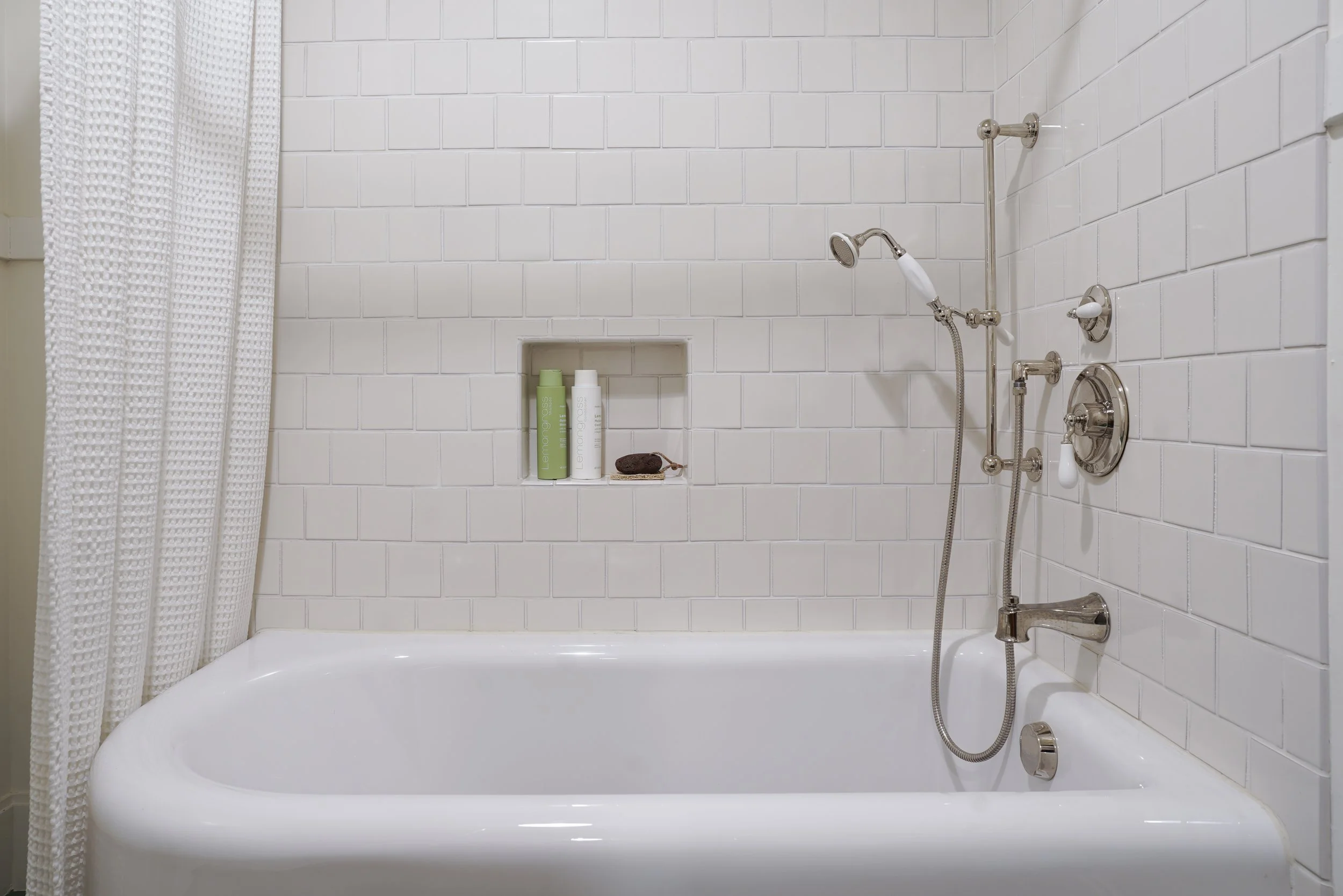 A white bathtub located against a tiled bathroom wall with a built-in shelf holding shampoo and conditioner bottles, and a brown washcloth, with a shower head and faucet on the right side.