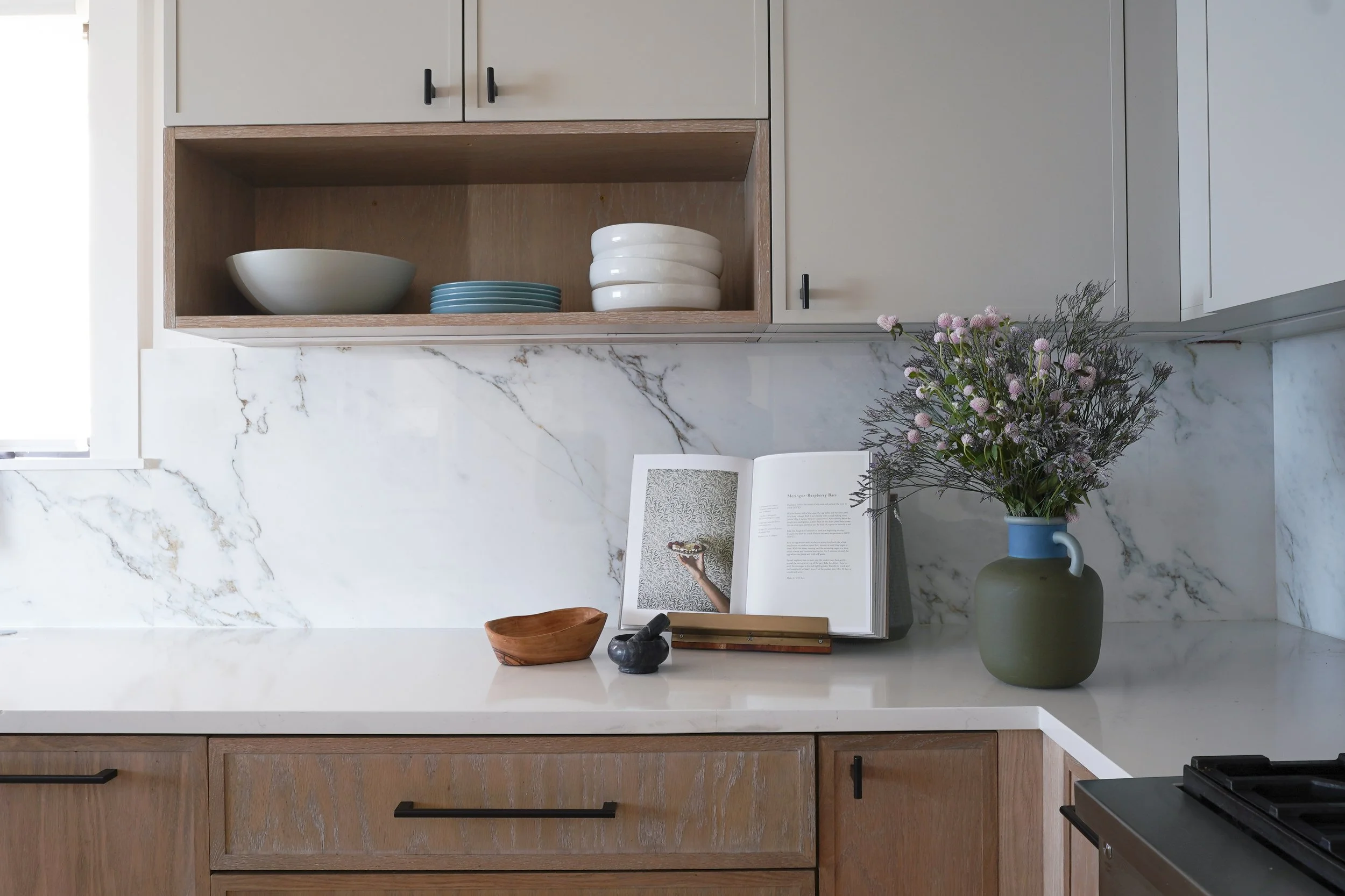 A kitchen countertop with an open cookbook, a green vase with pink and purple flowers, a small wooden bowl, and a black oval object. Above the countertop are cabinets and open shelves with white and blue bowls and plates, and a marble backsplash.