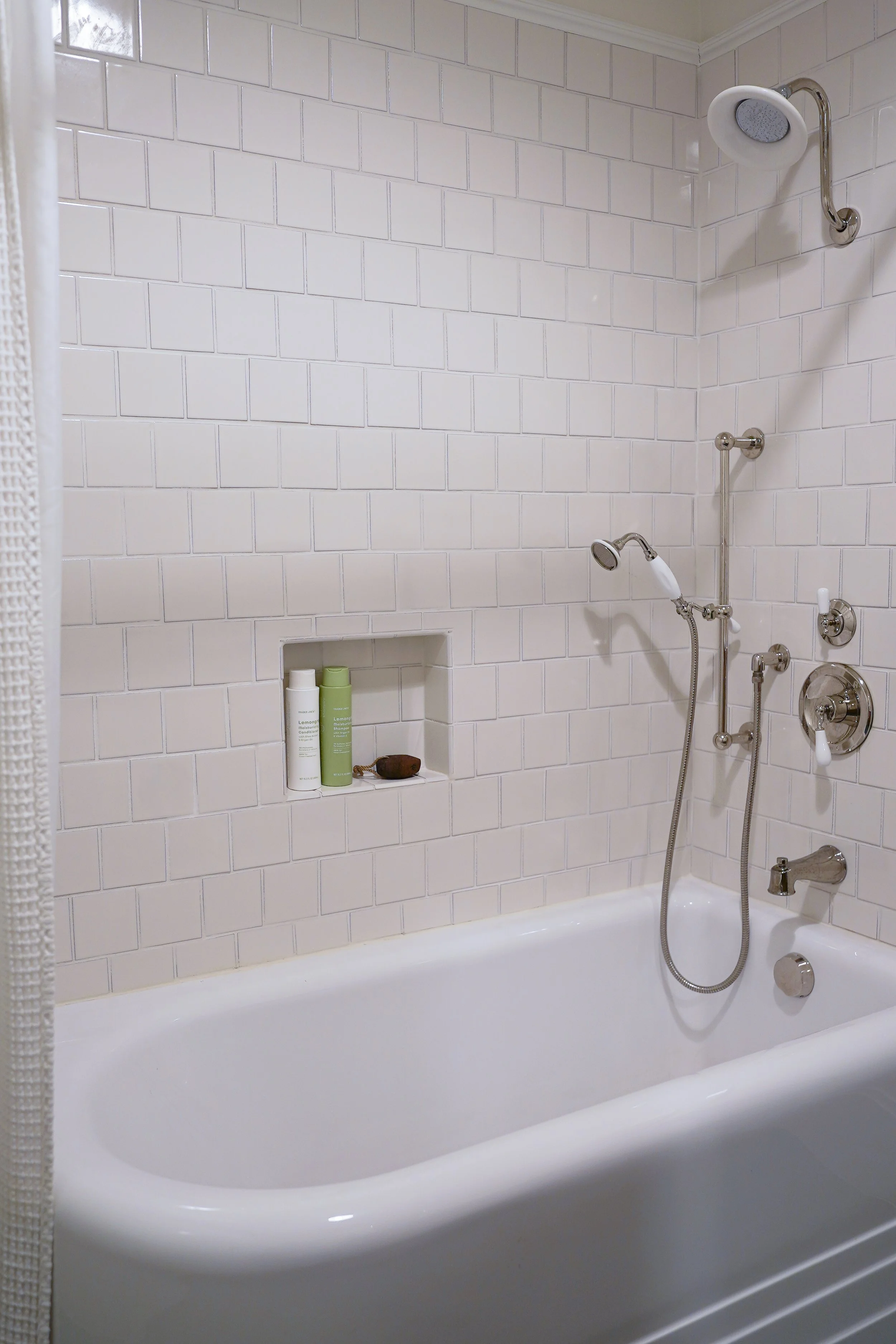A white bathtub in a bathroom with white subway tile walls. There are two shower heads and multiple pipes, one on a vertical bar. A small built-in shelf in the wall holds bottles and a stone object.