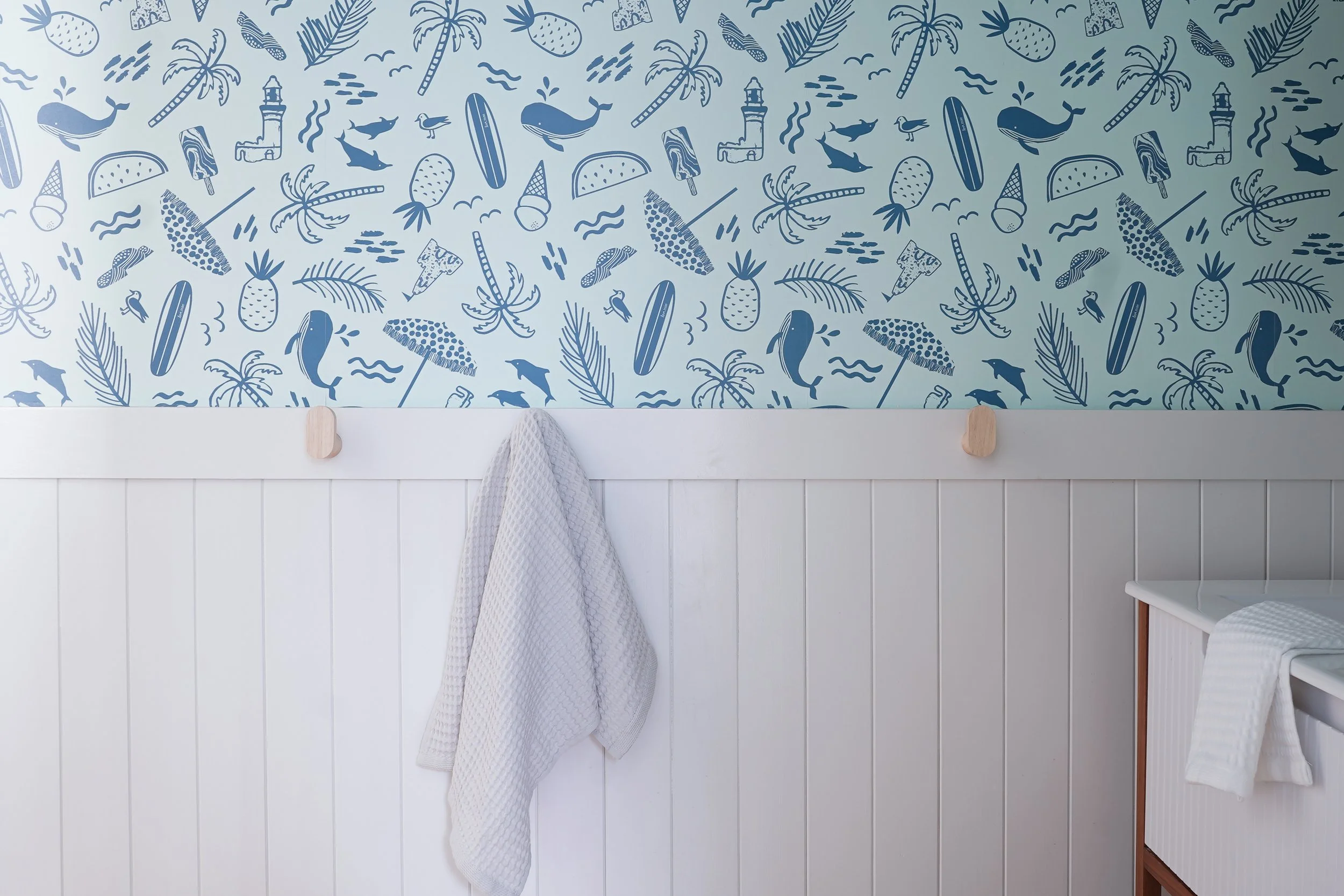 A white wall with vertical wooden paneling and nautical-themed wallpaper featuring whales, pineapples, palm trees, lighthouses, and surfboards in shades of blue. Two hooks hold a white towel, and part of a white chair with a textured cushion is visib