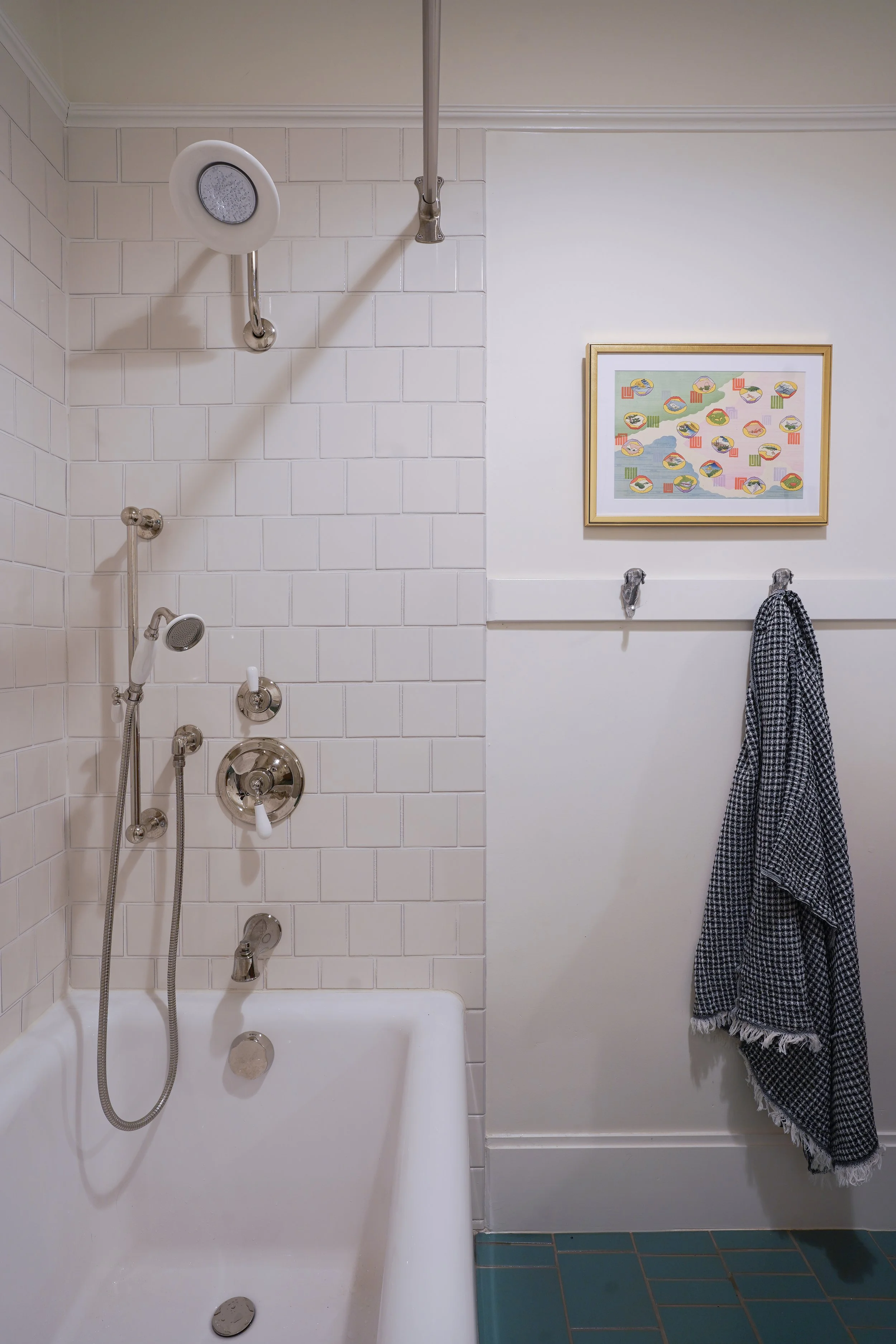 A bathroom with a shower and bathtub on the left, white tiled walls, and a framed colorful abstract art piece on the wall on the right, with a black and white checkered towel hanging from a hook.