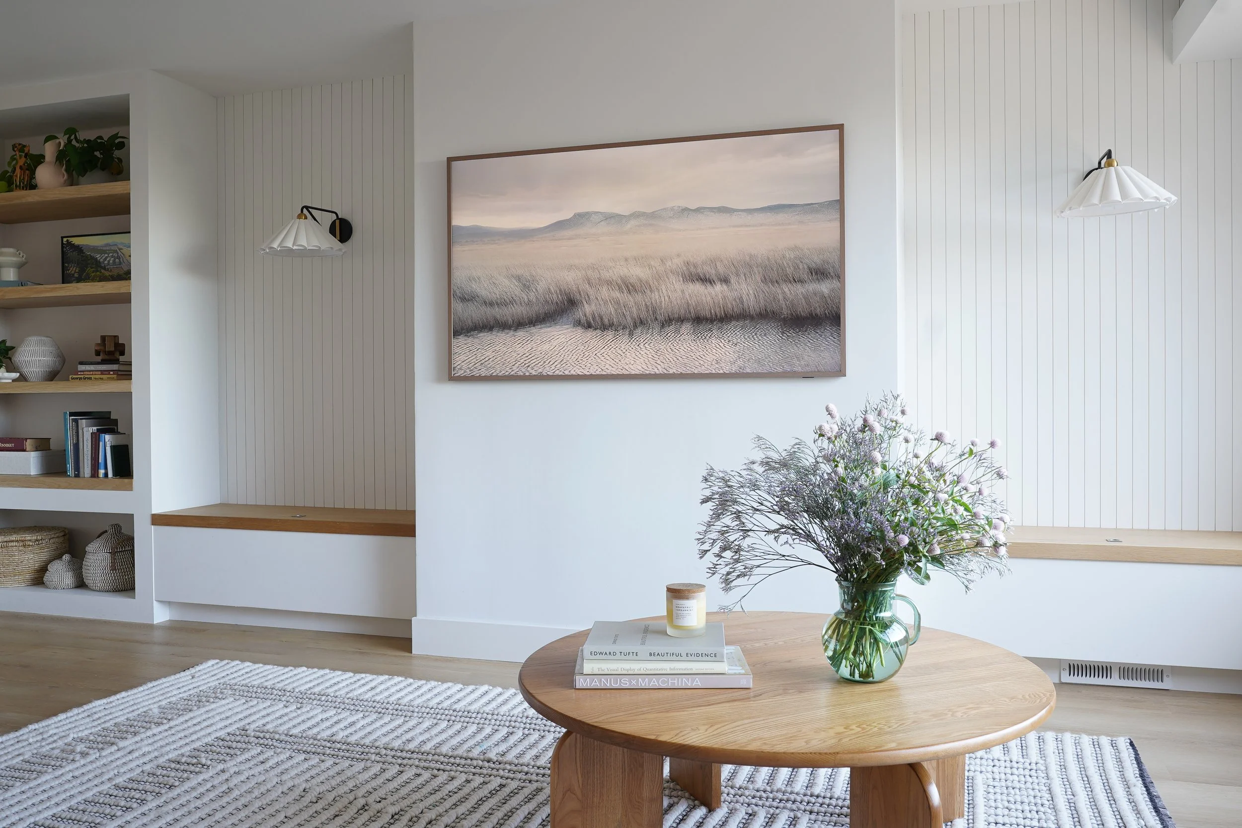 Living room with white walls and wooden accents, featuring abstract landscape artwork, a glass vase with purple flowers on a wooden table, and built-in shelves with decorative items.