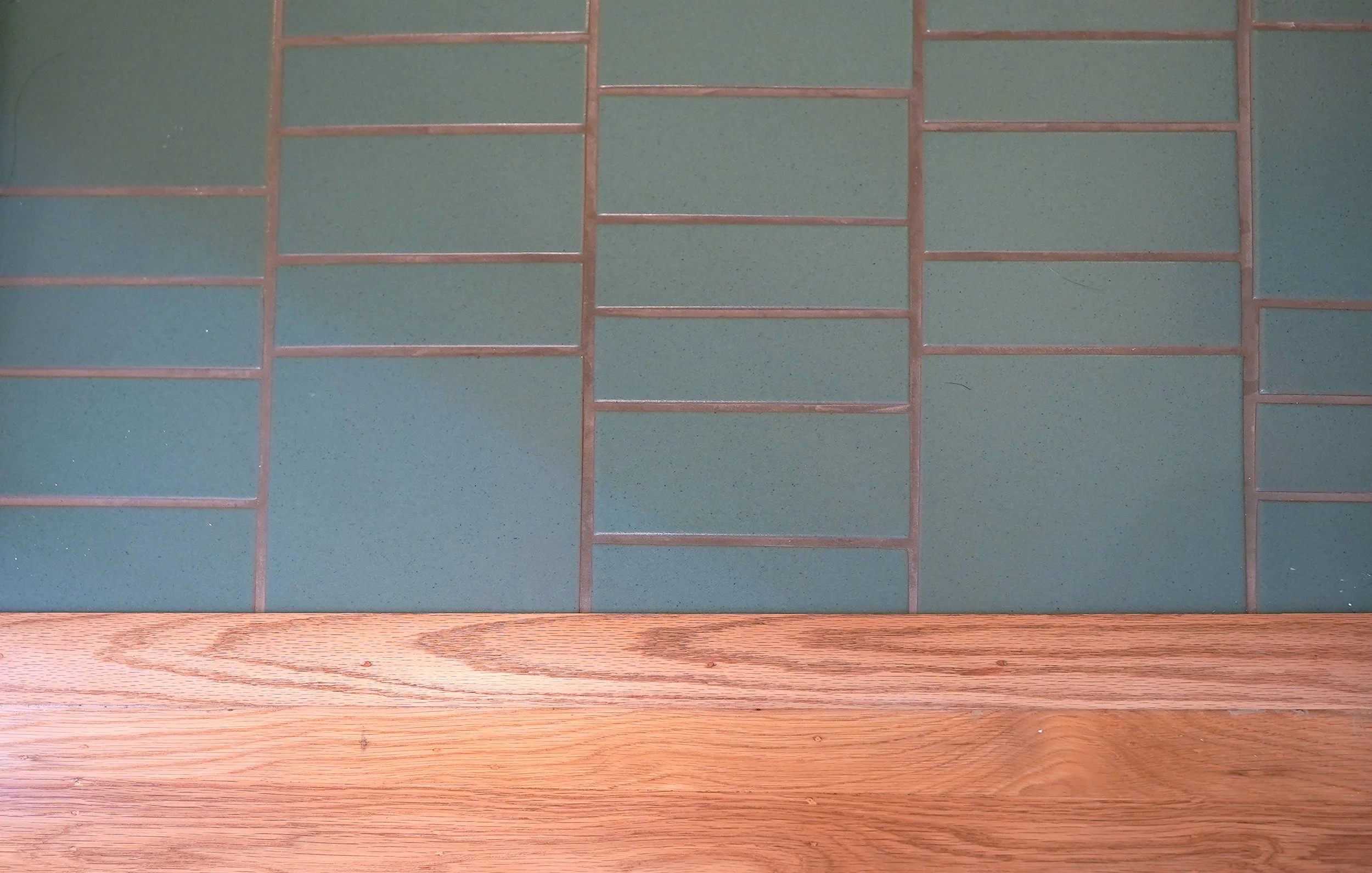 Close-up of green ceramic tiles with pink grout on a wall, with a wooden surface at the bottom.