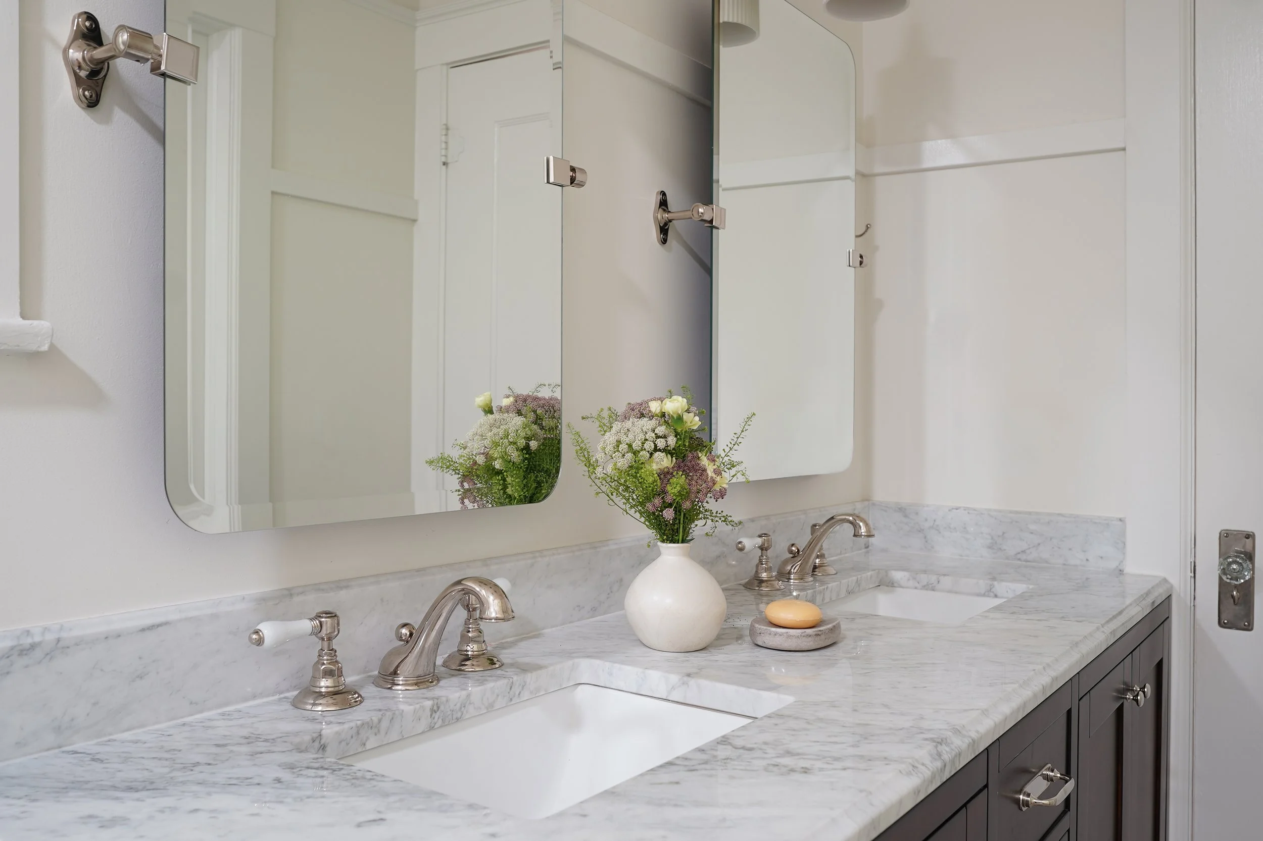 A bathroom sink with a marble countertop, two bronze faucets, and a large mirror above it. There is a white vase with flowers and some small stones on the counter and a reflection of a door and window in the mirror.