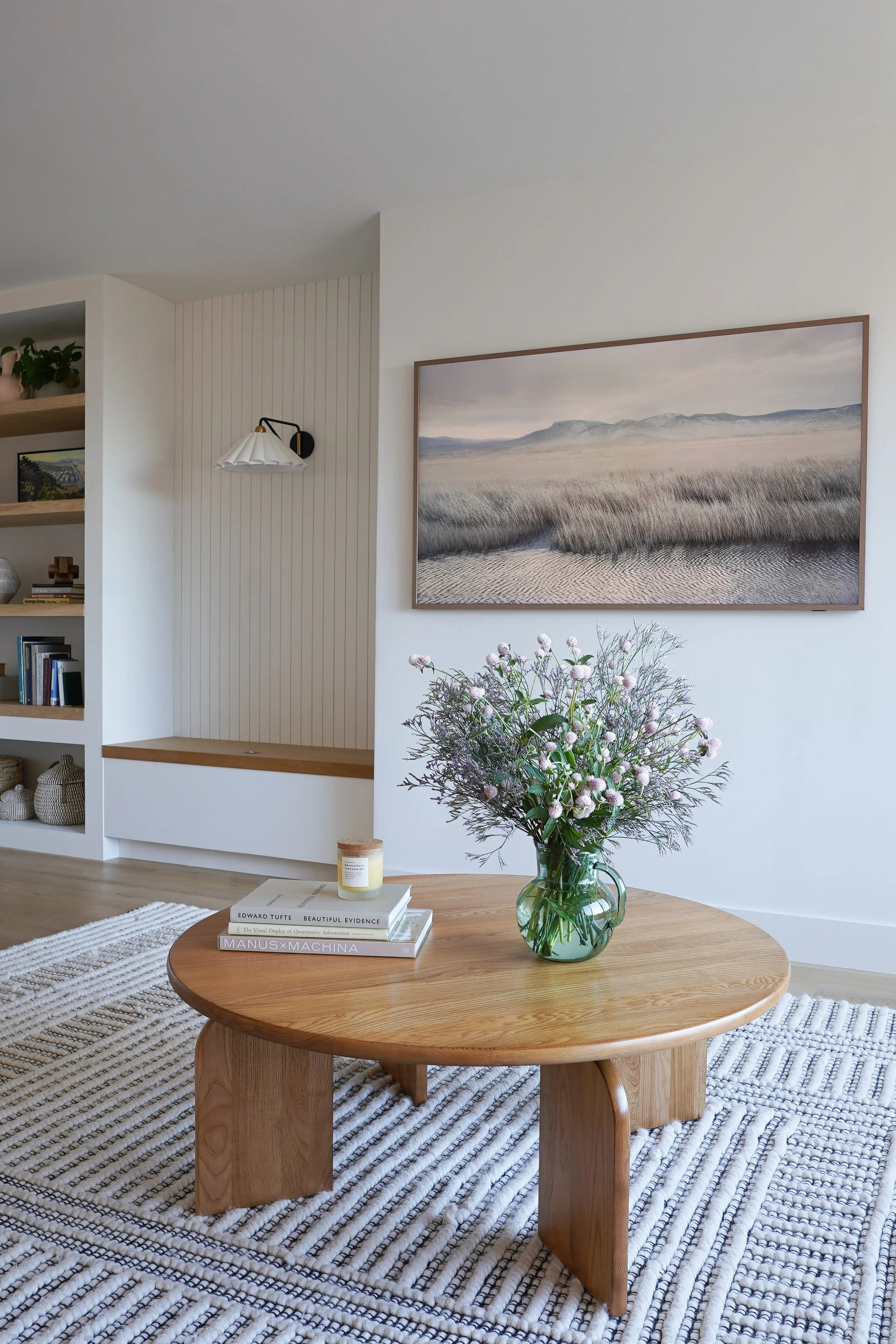 A round wooden coffee table with a glass pitcher of flowers, books, and a candle on top, placed on a striped rug in a minimalist living room with white walls, a large landscape painting, wall-mounted lamp, and built-in shelving.