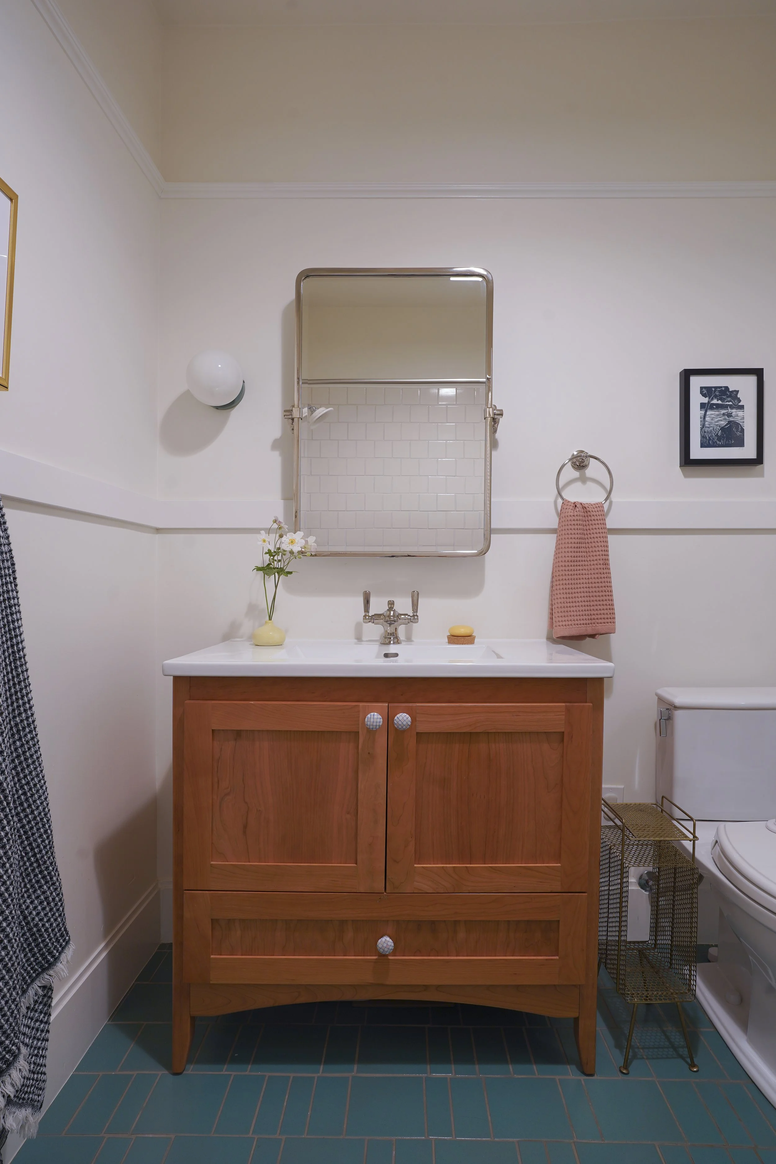 A bathroom vanity with a wooden cabinet and a white countertop. Above it, a rectangular mirror with a metal frame. To the right, a hand towel hanging on a ring, a framed picture of a lake scene on the wall, and a small white bowl with a soap dish. To