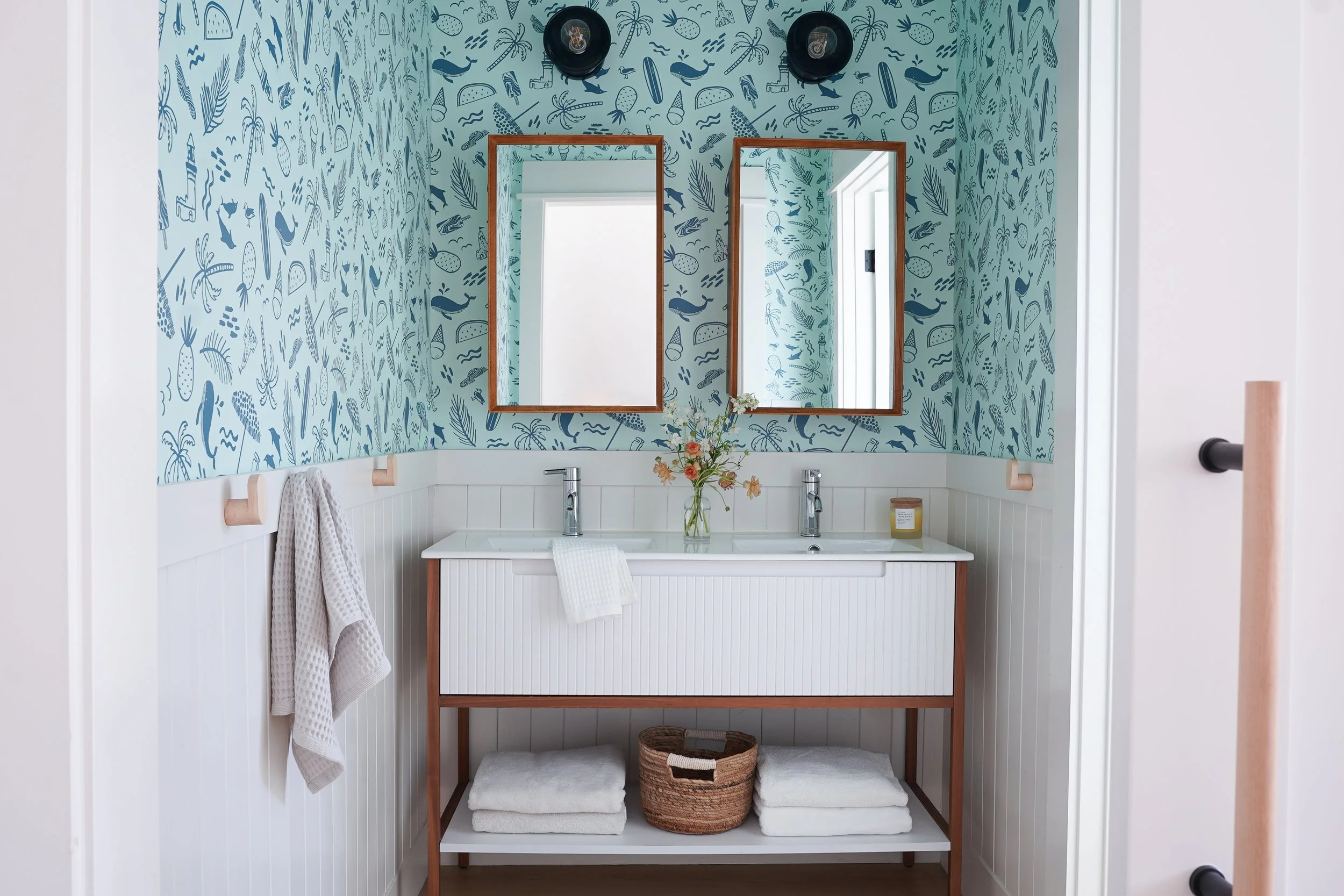 A bathroom vanity area with a shower curtain, two mirrors, a flower arrangement, towels, and baskets, against a wall with blue nautical-themed wallpaper featuring whales, fish, and marine elements.