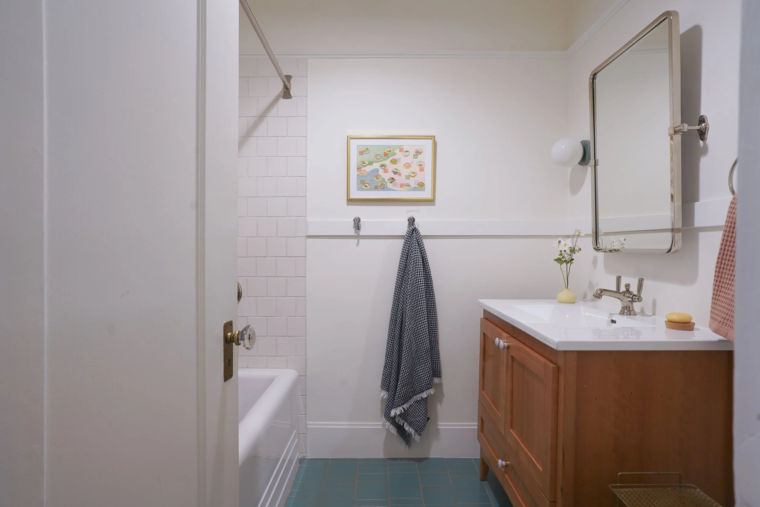 A small bathroom with a white bathtub on the left, a wooden vanity with a white sink and a large mirror on the right, and a framed colorful artwork on the wall. There are towels hanging and a small plant on the vanity.
