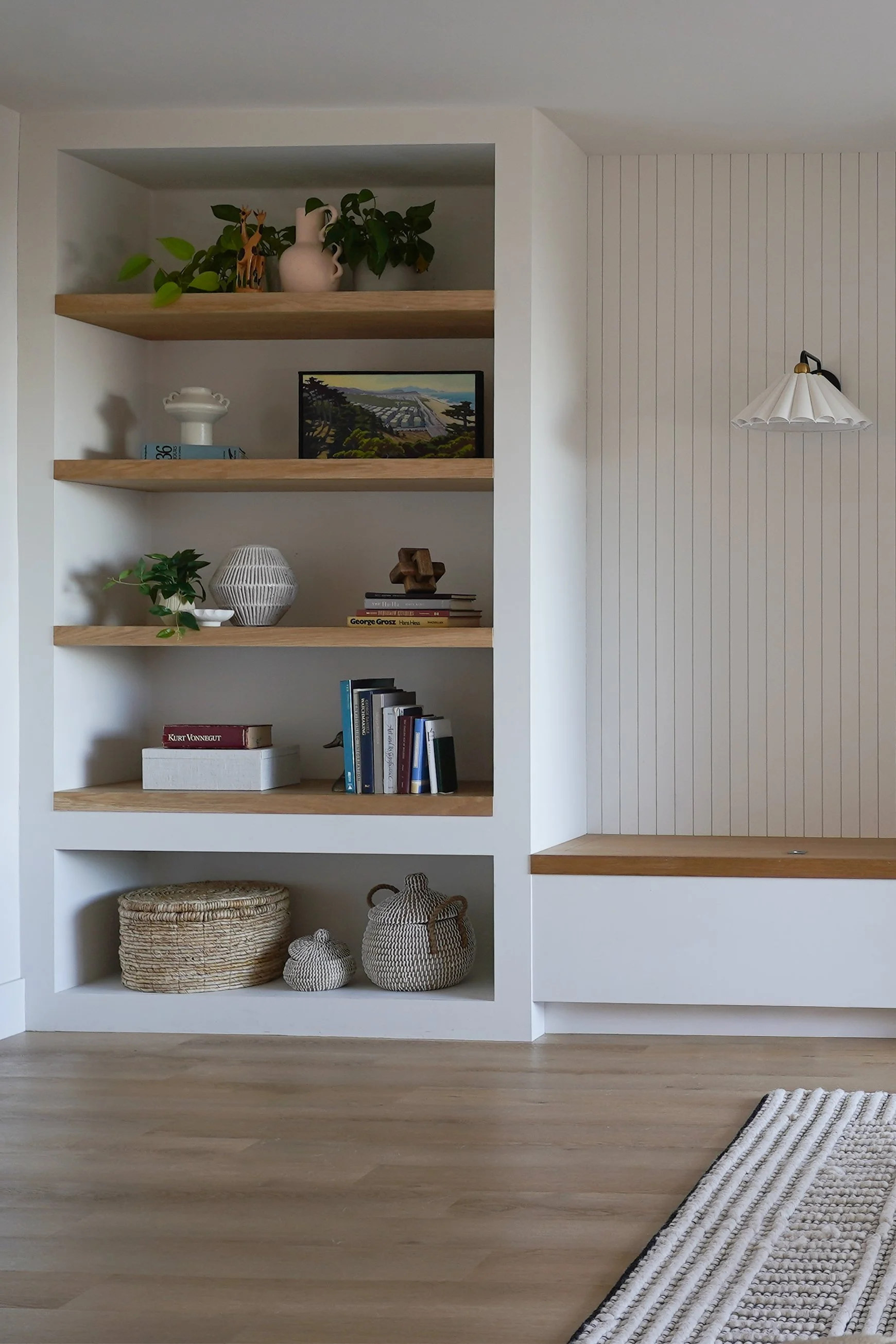 A modern, minimalist bookshelf with decorative items including vases, books, and plants, against a white wall with wooden accents.