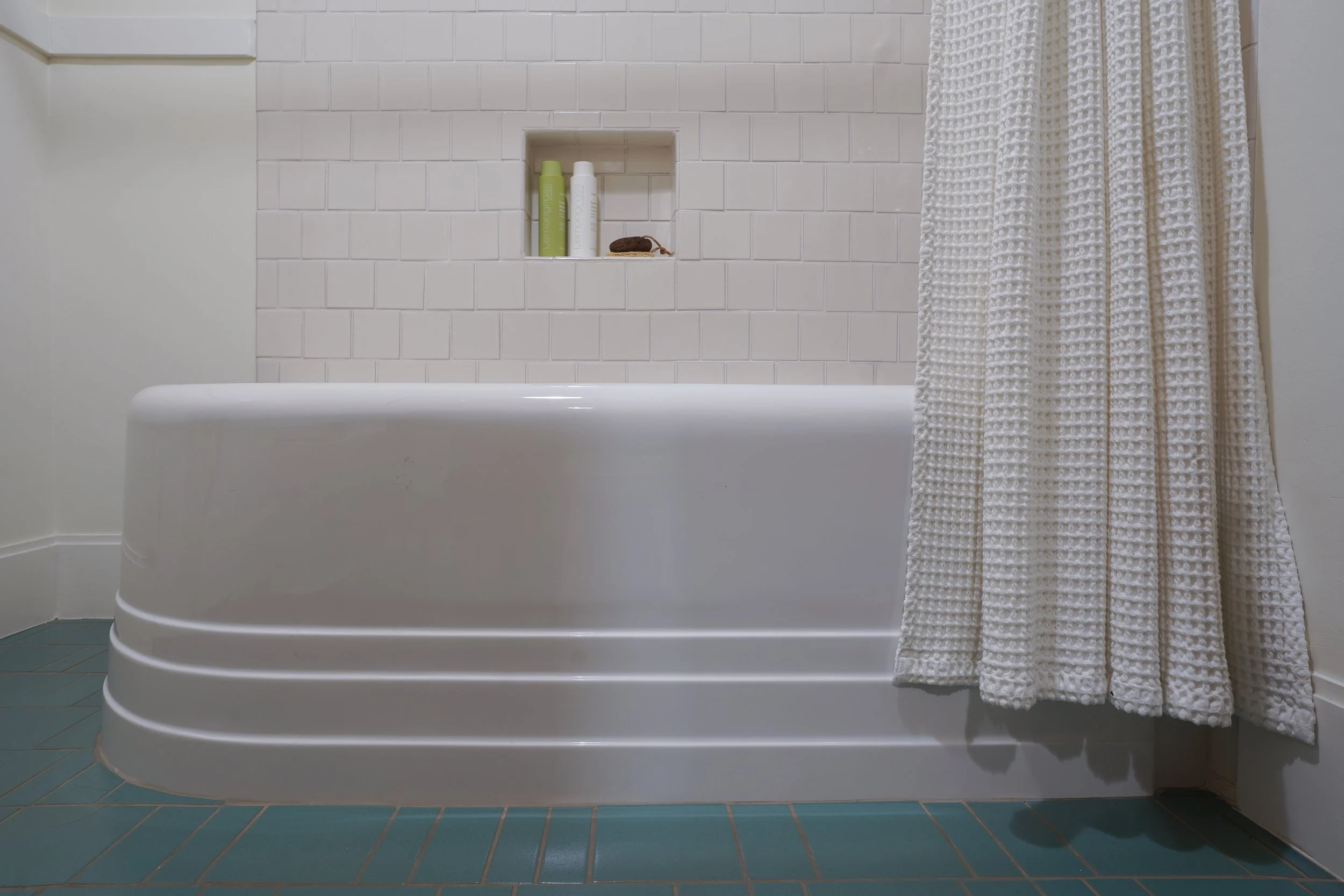 View of a bathroom corner with a white bathtub, textured white shower curtain, built-in wall niche with shampoo bottles, and teal floor tiles.