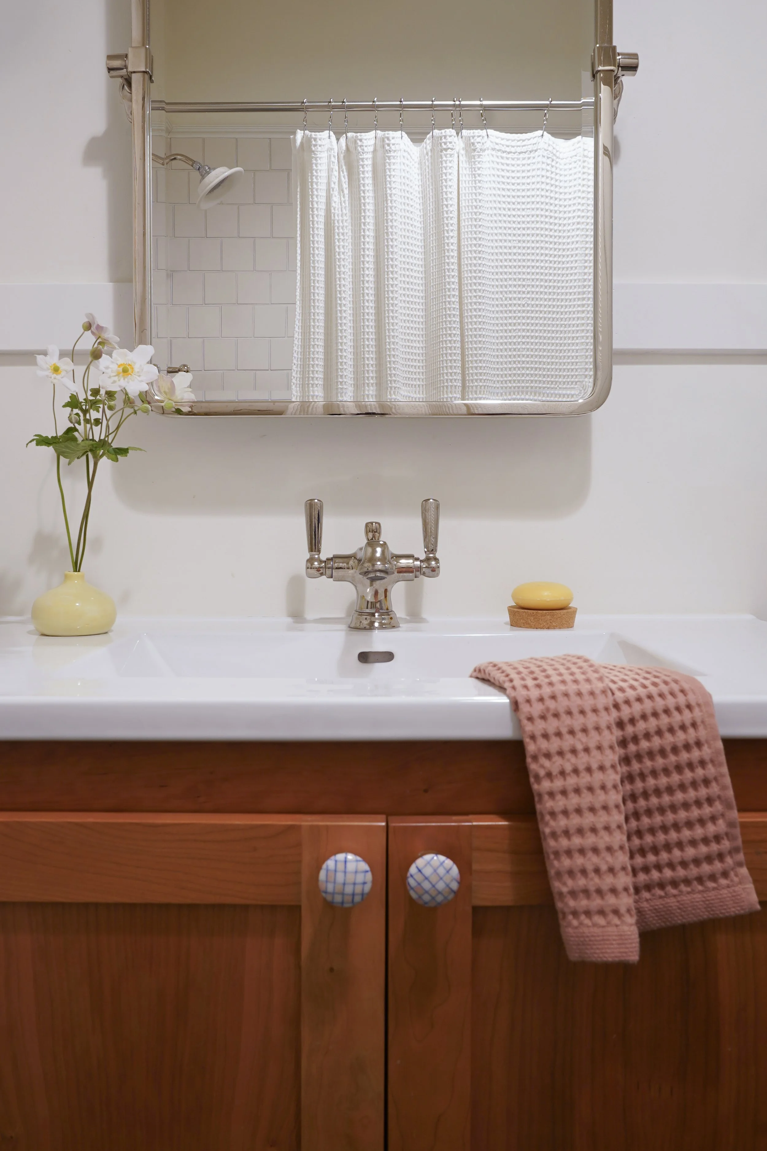Bathroom sink with a wooden cabinet, pink towel, soap on a cork coaster, a yellow vase with white flowers, a mirror with white shower curtain, showerhead, white tiled wall.