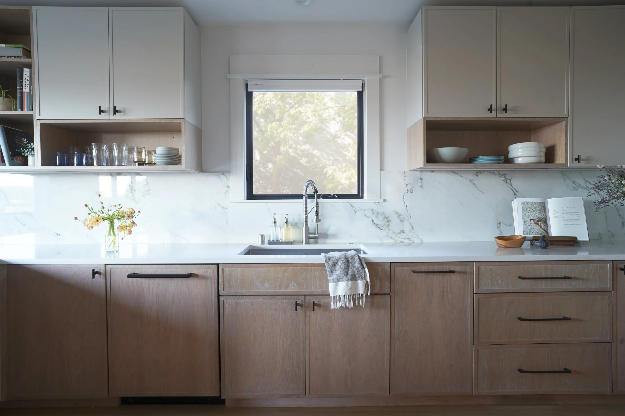 Modern kitchen with wooden lower cabinets, white upper cabinets, a marble backsplash, a window with a view of greenery, and various kitchen items including dishes, glasses, a book, and a vase with flowers.