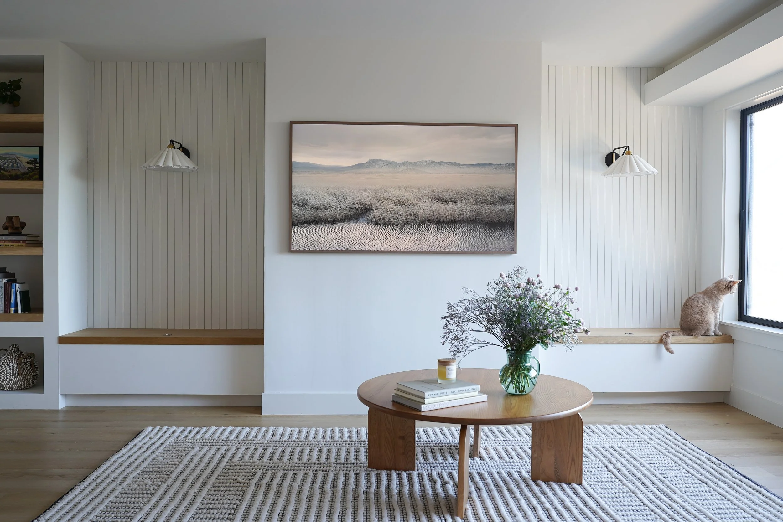 Living room with white shiplap walls, a large landscape painting, two wall-mounted lamps, a round wooden coffee table with books and a vase of flowers, a median striped rug, and a window ledge with a cat sitting and looking outside.