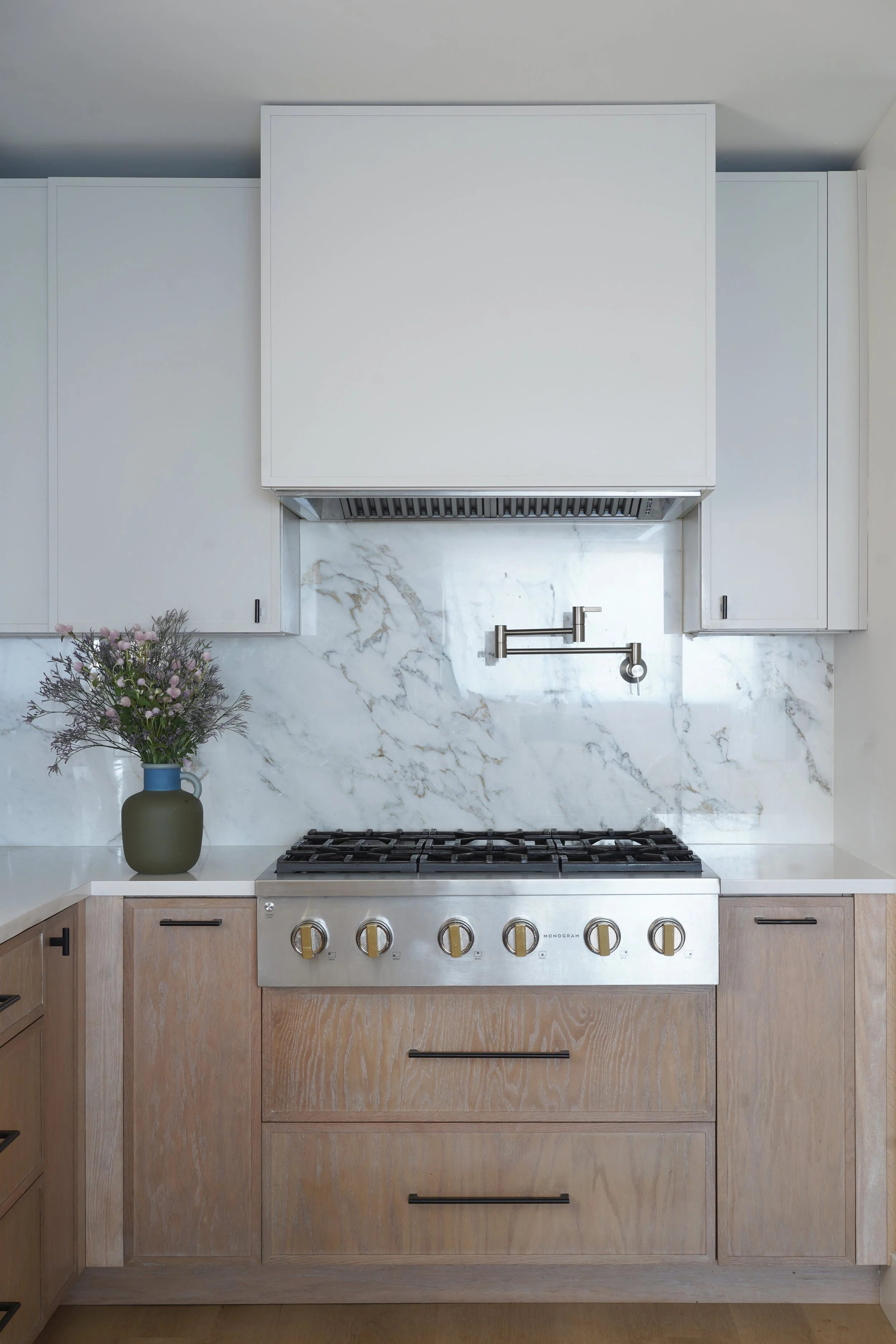 Modern kitchen with white marble backsplash, a stainless steel gas stove with gold knobs, light wood cabinetry, a silver range hood, and a vase with purple flowers on the countertop.