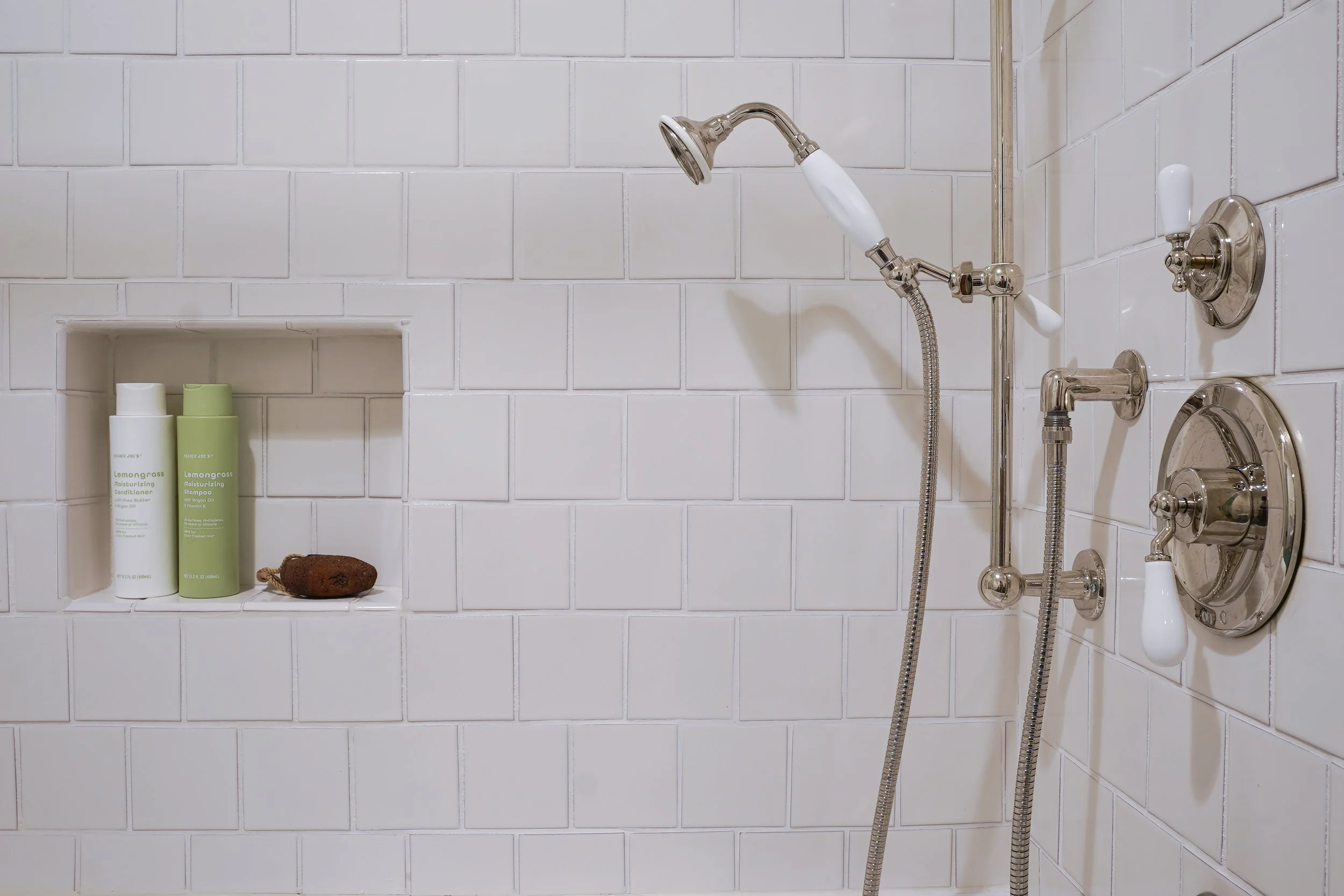 Shower with white subway tiles, built-in niche with two containers of hair care products, a loofah, and a handheld showerhead.