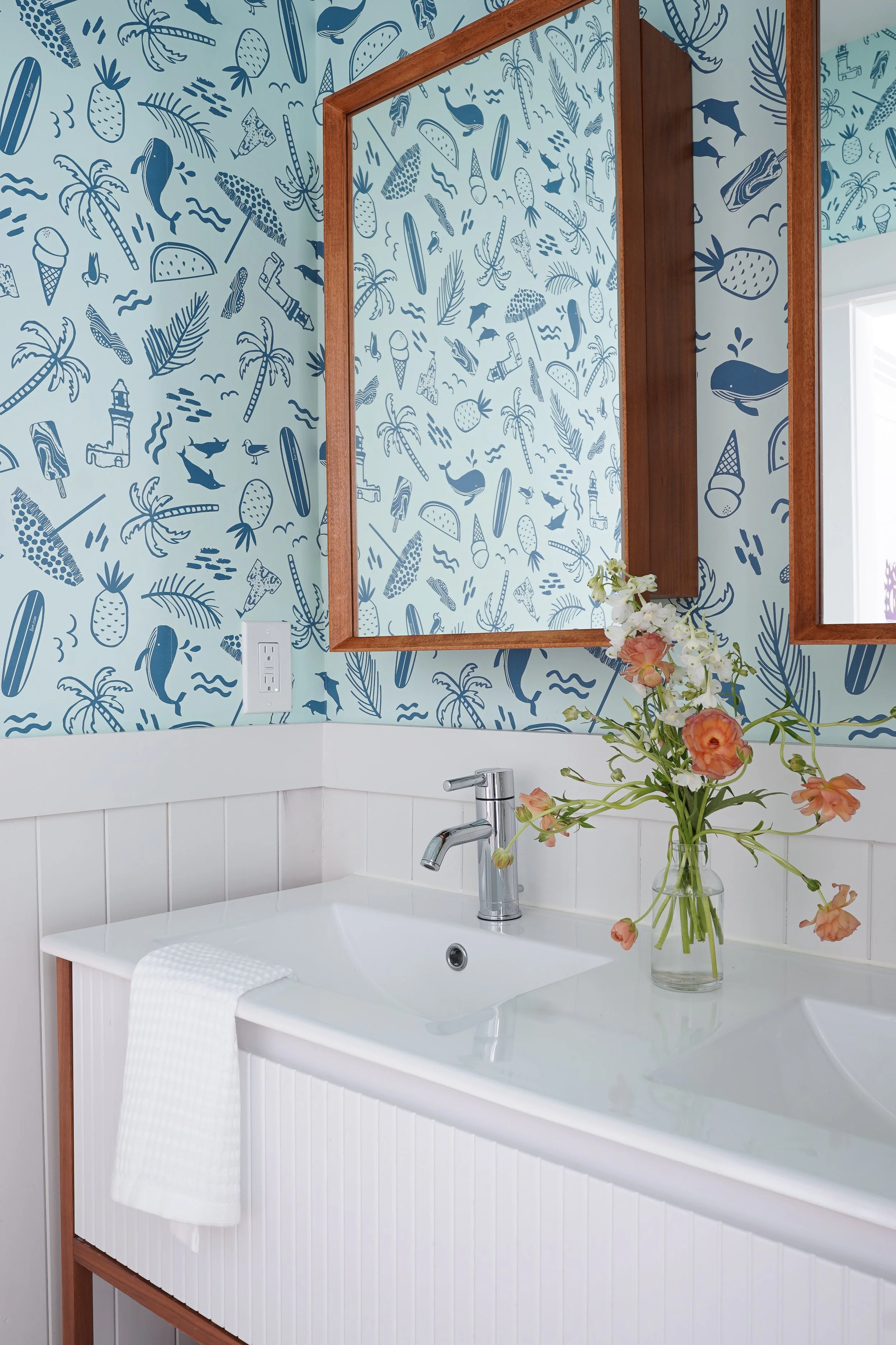 A bathroom with a white sink, a silver faucet, and a glass vase with peach and white flowers on the countertop. The wall has tropical-themed wallpaper with pineapples, whales, dolphins, palms, and ice cream cones in blue and white. A large rectangula