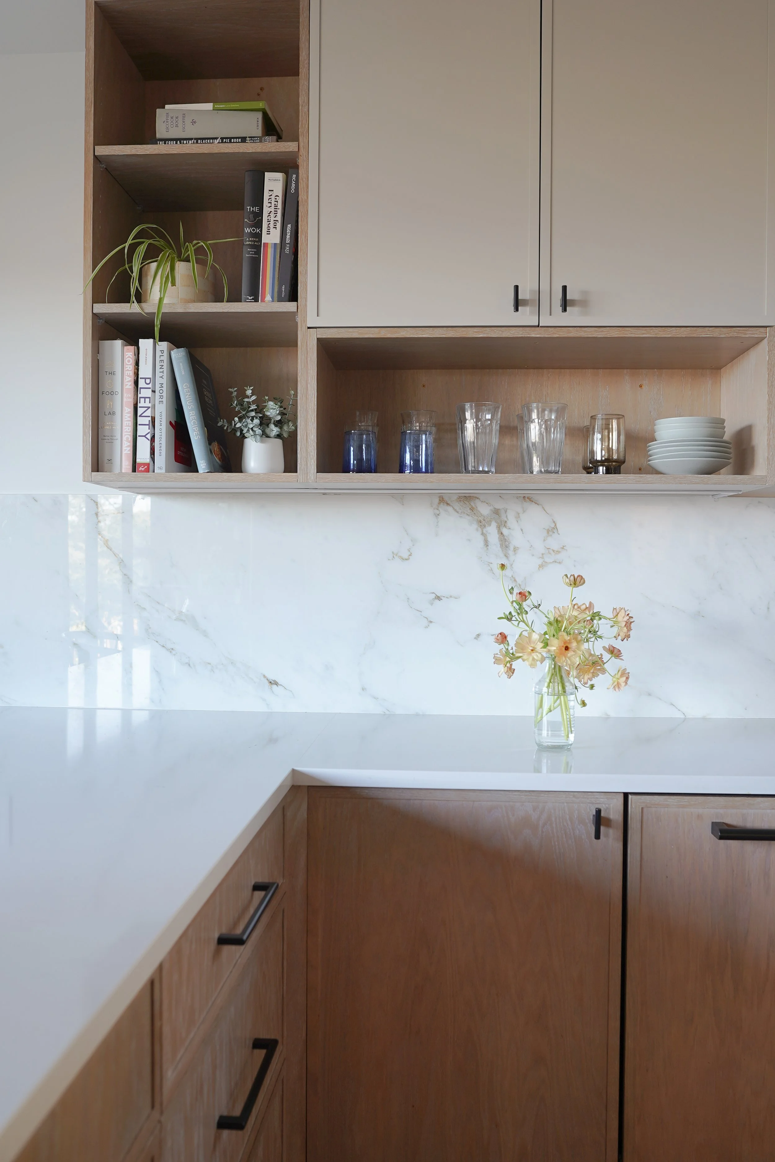 Open wooden kitchen cabinets with books, a potted plant, glasses, bowls, and flowers on a marble countertop.