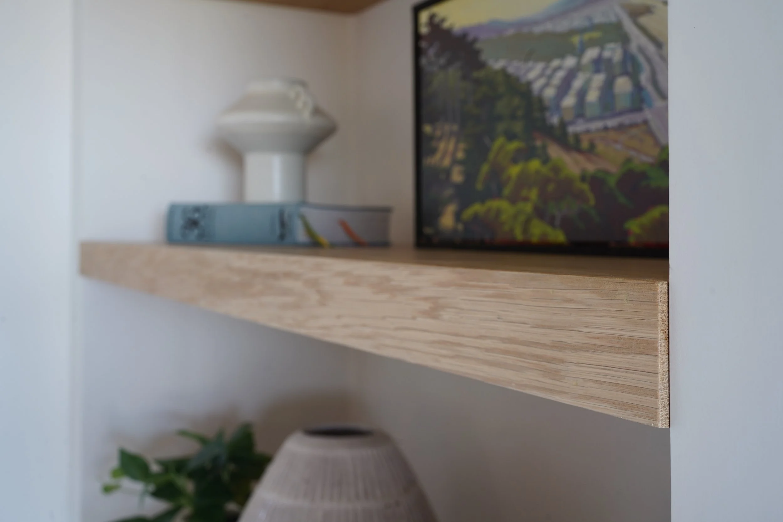 A close-up of a wooden shelf with a white ceramic vase, a rectangular box with a landscape painting featuring houses and greenery, and a green plant partially visible at the bottom.