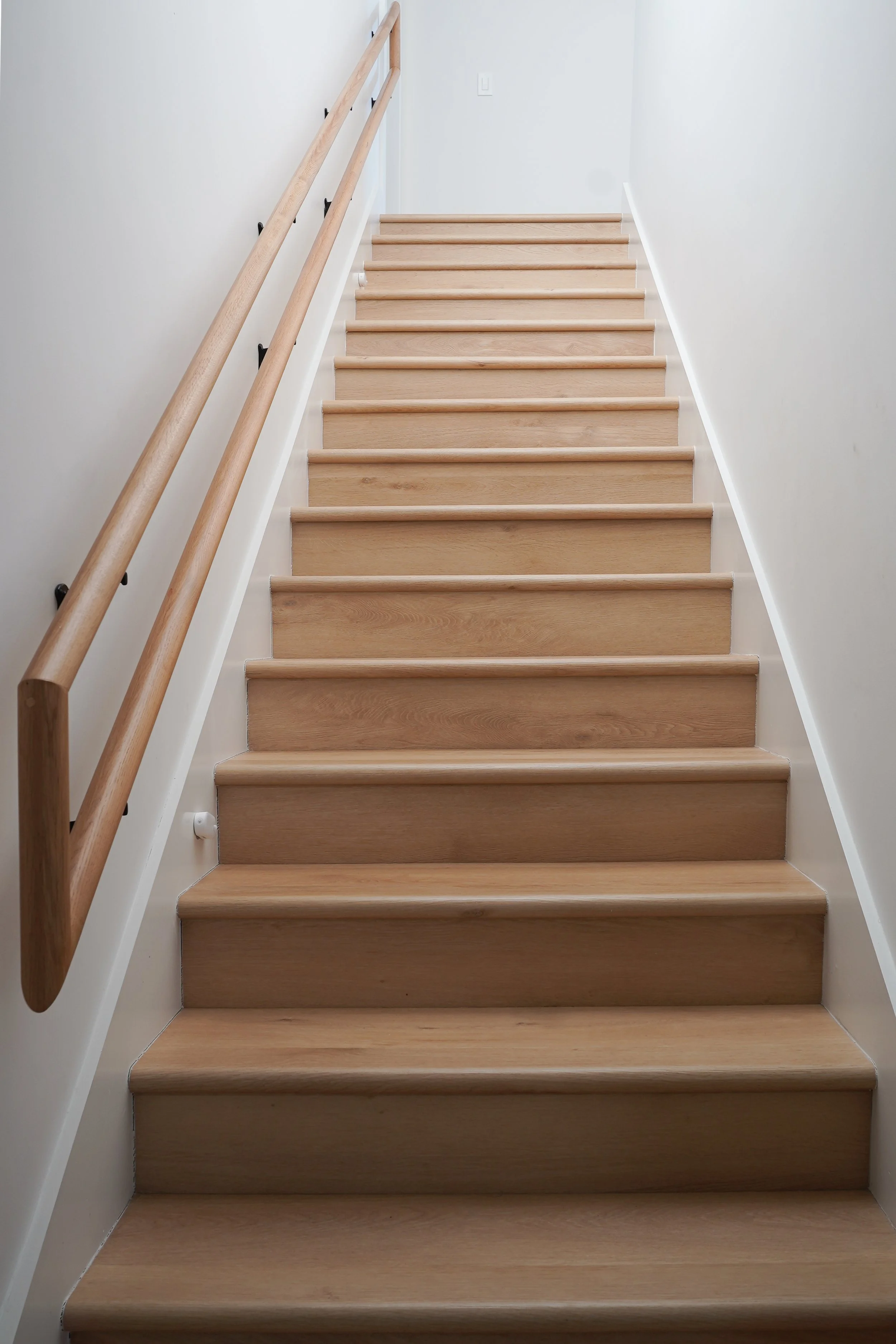 Wooden staircase with a handrail on the left side, white walls on either side, and a light switch at the top.