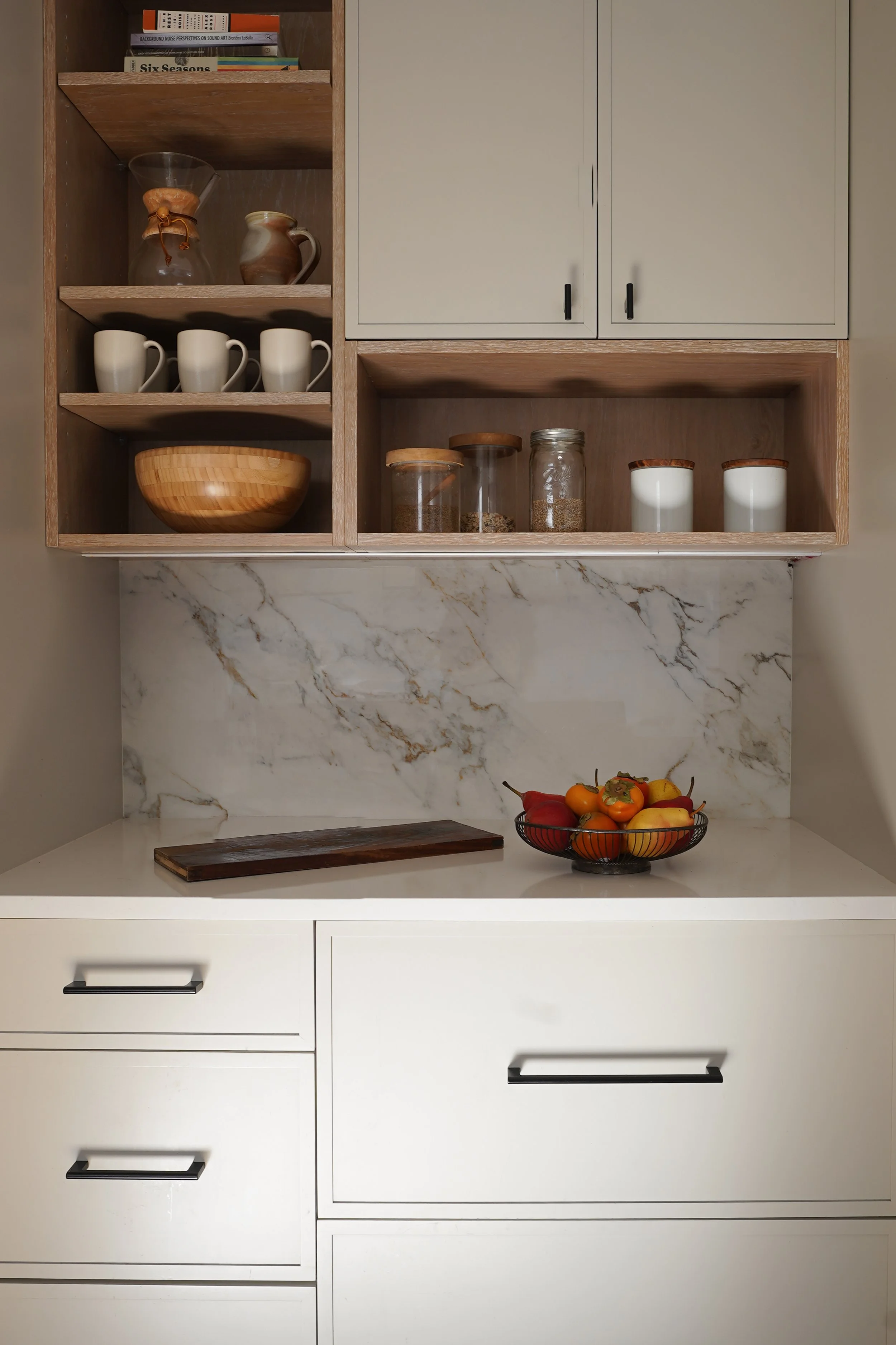 Kitchen cabinetry with open shelves holding books, a glass coffee dripper, mugs, a wooden bowl, and jars of grains, with a white marble backsplash and a bowl of apples and peaches on the counter.