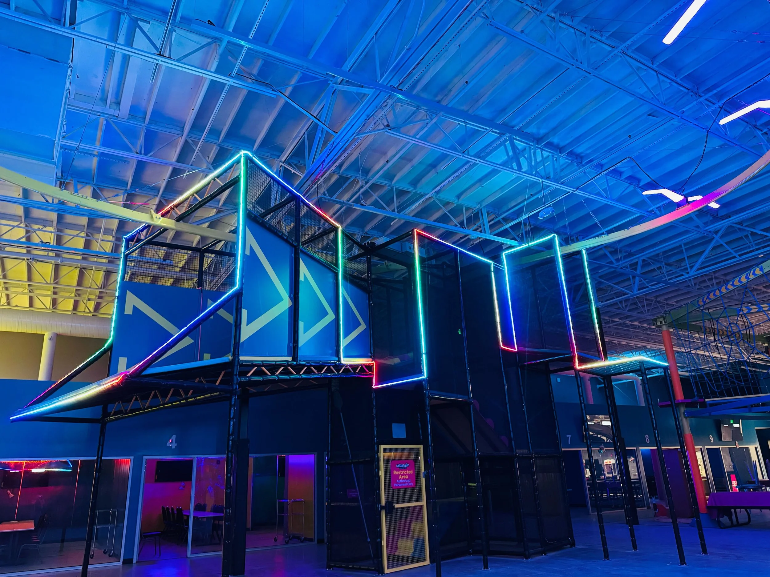 Indoor playground with play structure outlined in colorful neon lights, including slides and climbing areas, inside a spacious room with a high ceiling and exposed metal beams.
