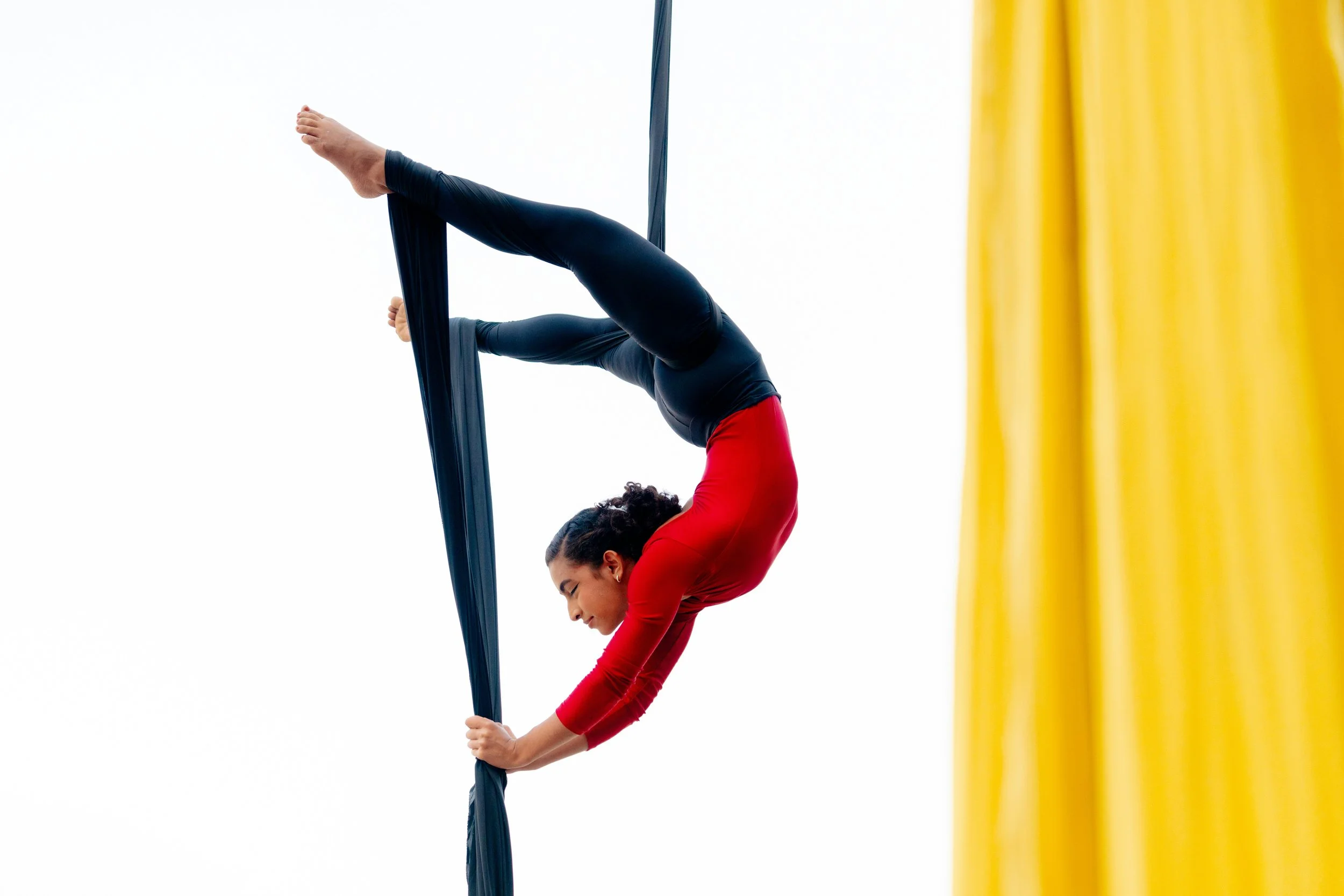 A woman in a red top performing aerial silk acrobatics against a white background, partially obscured by a yellow curtain on the right side.