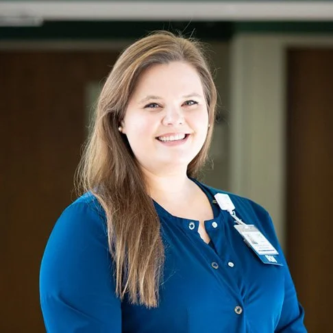 A smiling woman in a blue uniform with a name badge at a hospital or medical facility.