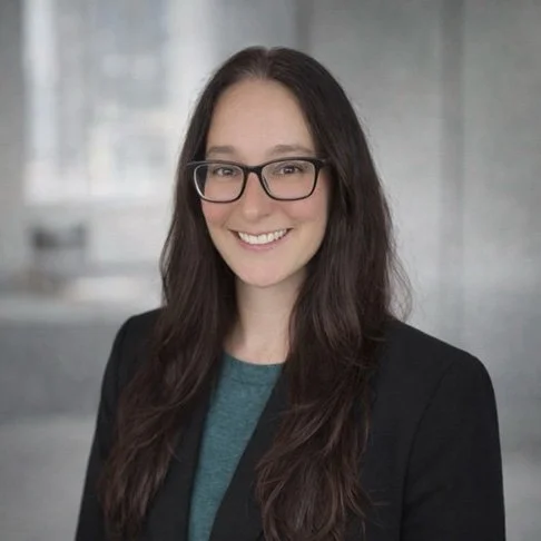 A woman with long dark hair, glasses, and a black blazer smiling in a professional setting.