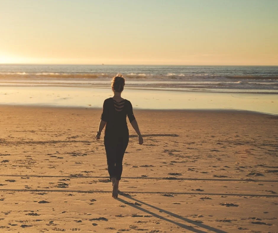 A woman is walking on a sandy beach at sunset, facing the ocean with waves, with her shadow extending behind her.