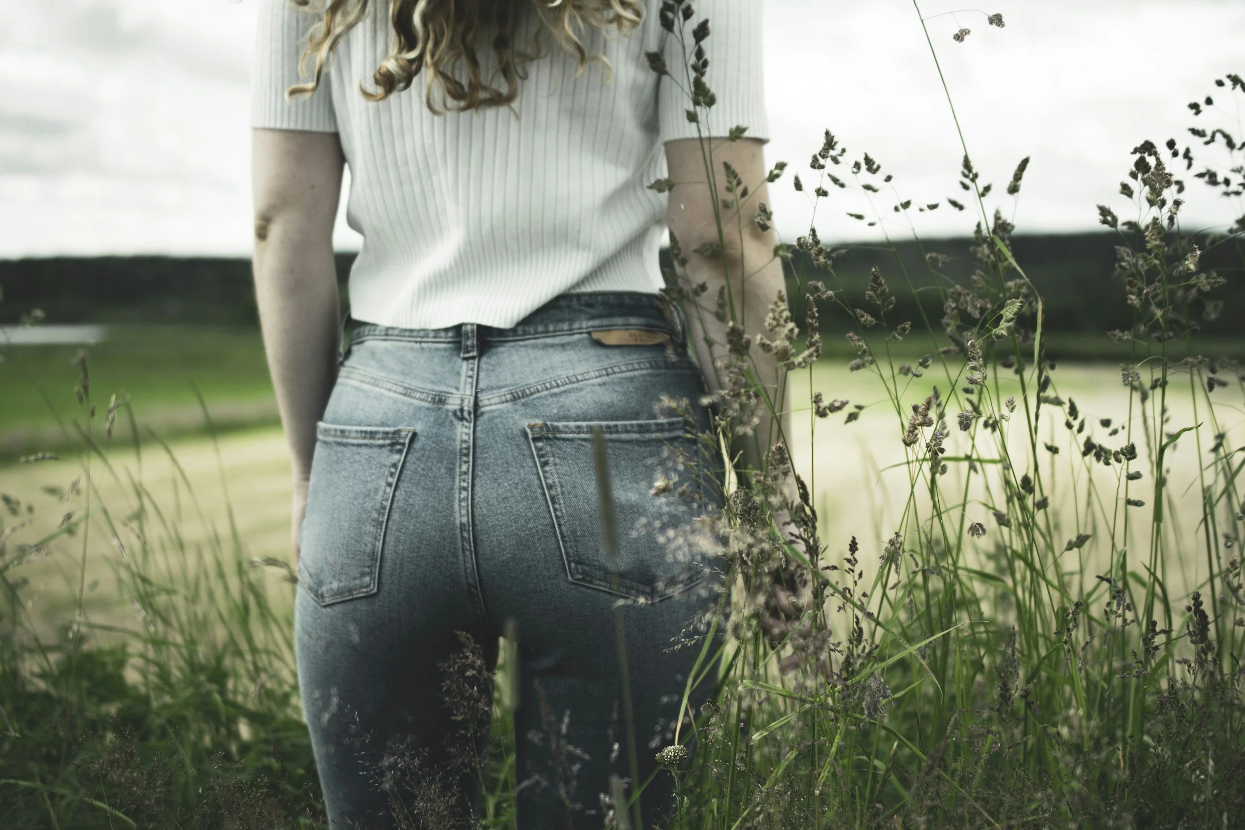 A woman standing in a field with tall grass, seen from behind, wearing a white ribbed top and high-waisted blue jeans. She has long, wavy hair.