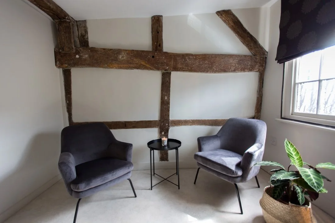 A cozy room featuring two gray armchairs, a small round black side table with a candle, a potted plant, exposed wooden beams on the wall, and a window with dark curtains allowing natural light.