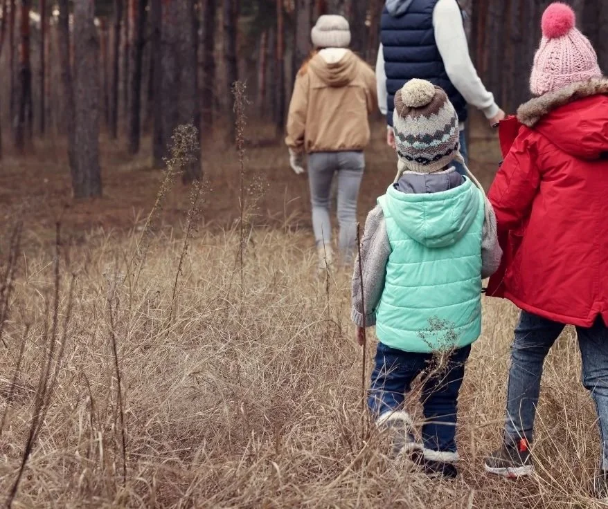 A group of children and an adult walking through a brown, grassy field in a forested area during fall or winter, all dressed in warm clothing and winter hats.