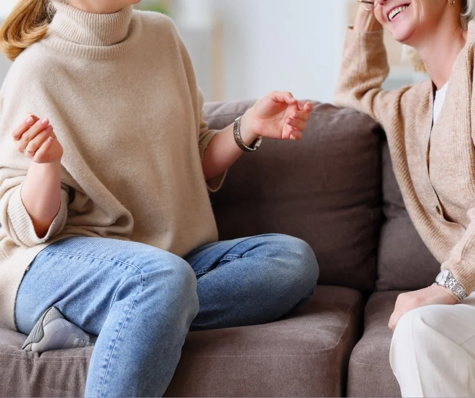 Two women sitting on a couch engaging in conversation, smiling, with one woman gesturing with her hand.