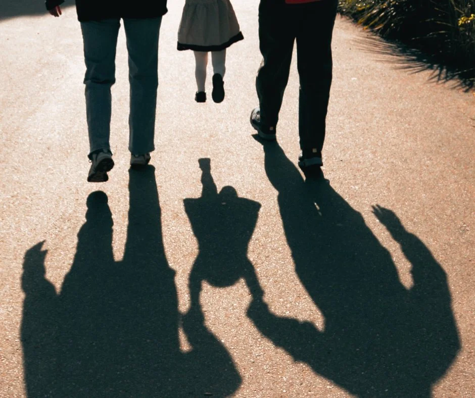 Photo of a person and a child walking on a sidewalk, with their shadows cast on the ground.