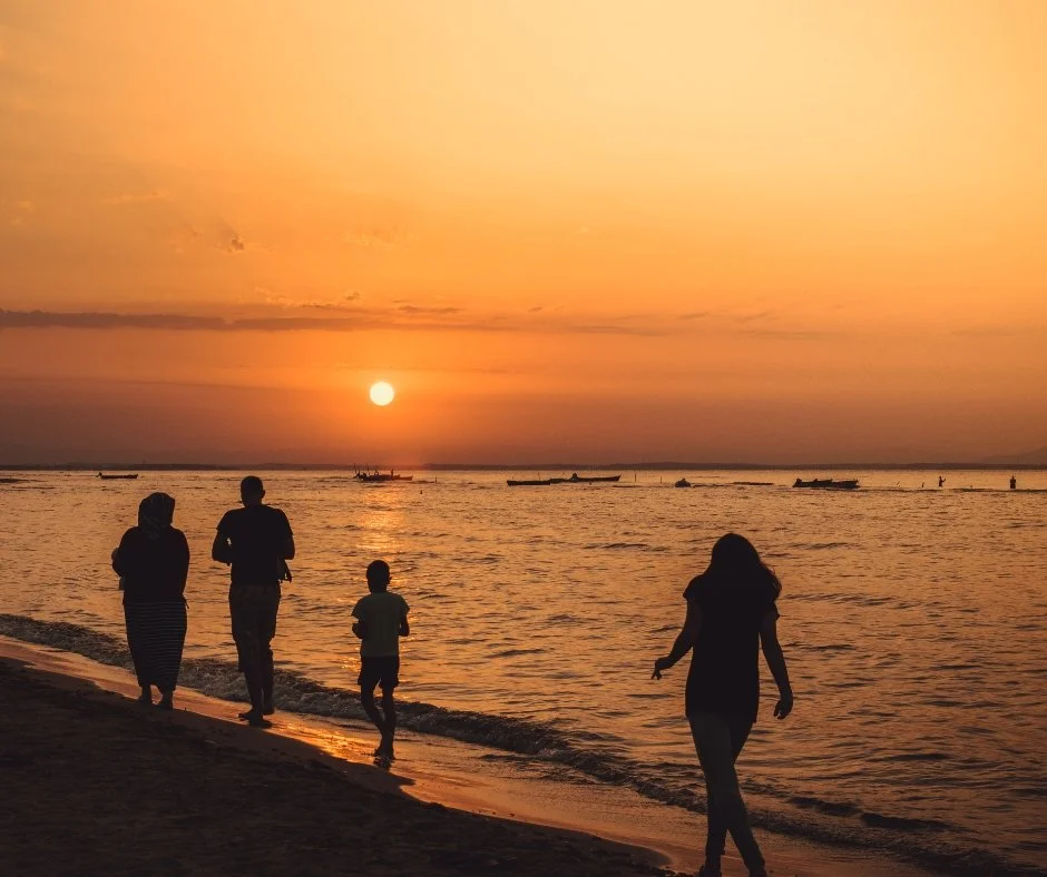 People walking along a beach during sunset with boats in the water.