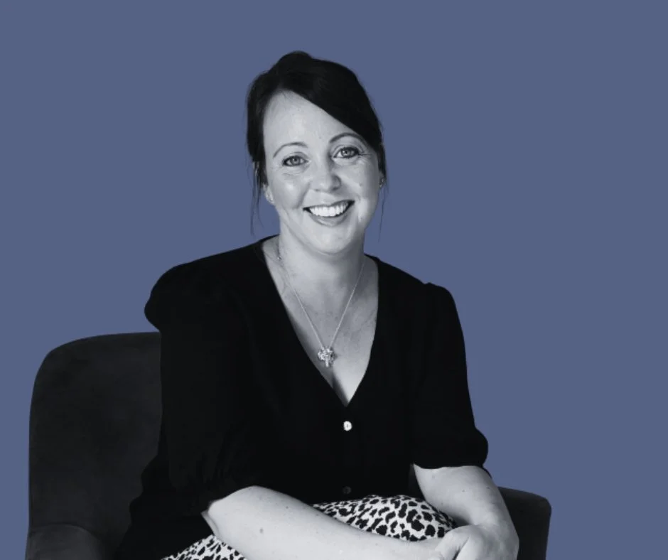 A woman with dark hair in a ponytail, smiling, wearing a black top, sitting on a chair against a blue background.