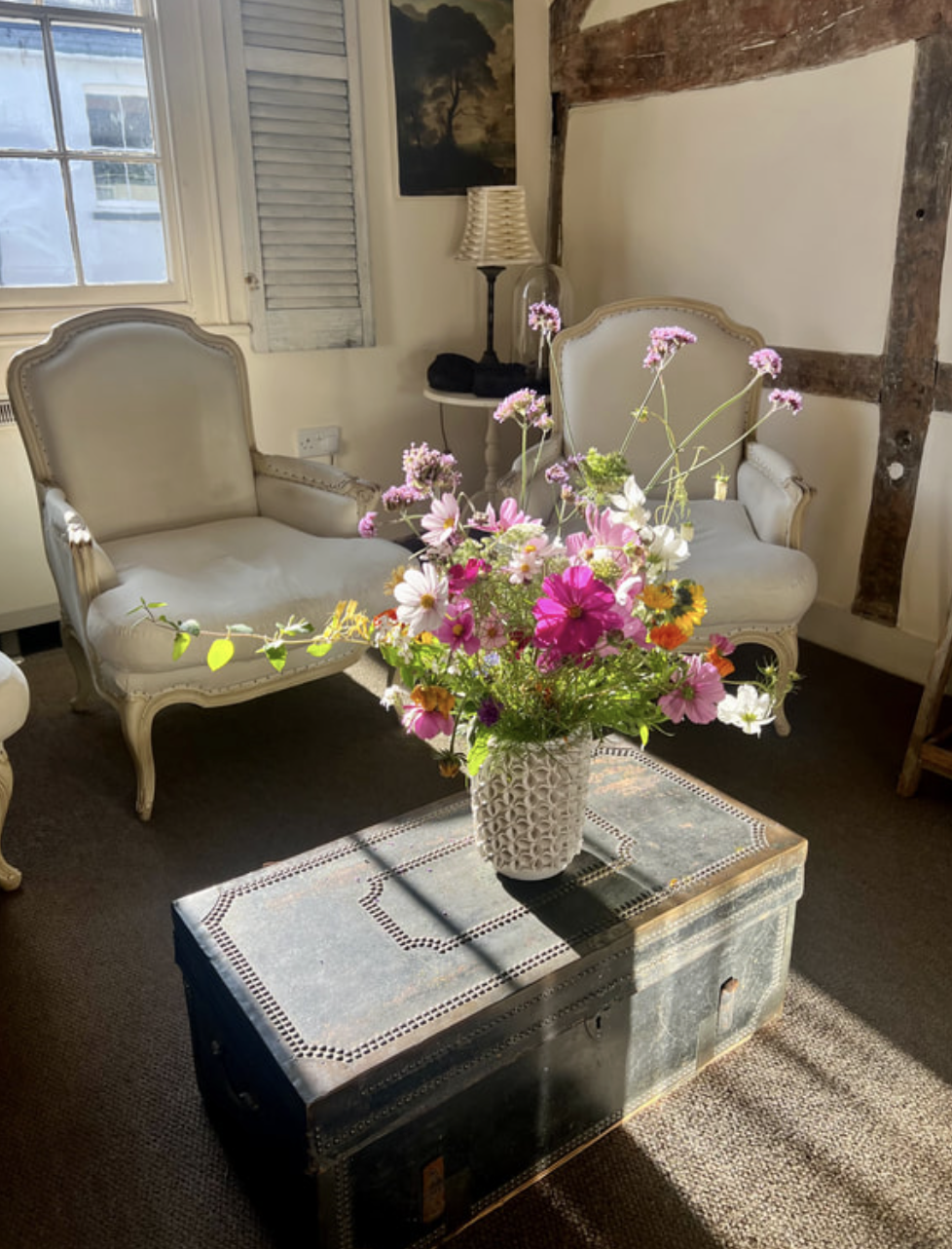 Pure Therapy room in Alton High street - with two vintage-style cream-colored sofas, a floral arrangement on a grey chest in the center, a window with light blue shutters.
