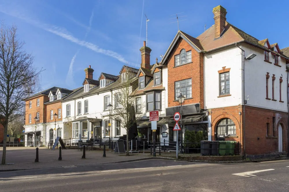 Street view of Alton Town centre - a row of multi-story buildings with mixed brick and white facades, some with gabled roofs and dormer windows, under a blue sky.