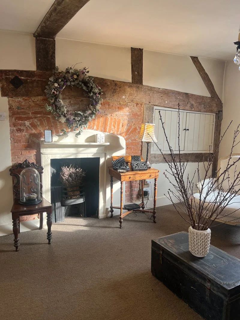 Living room with brick fireplace, floral wreath above, wooden beams on ceiling, small side tables with lamps, and a large vase with branches, in an rustic style.