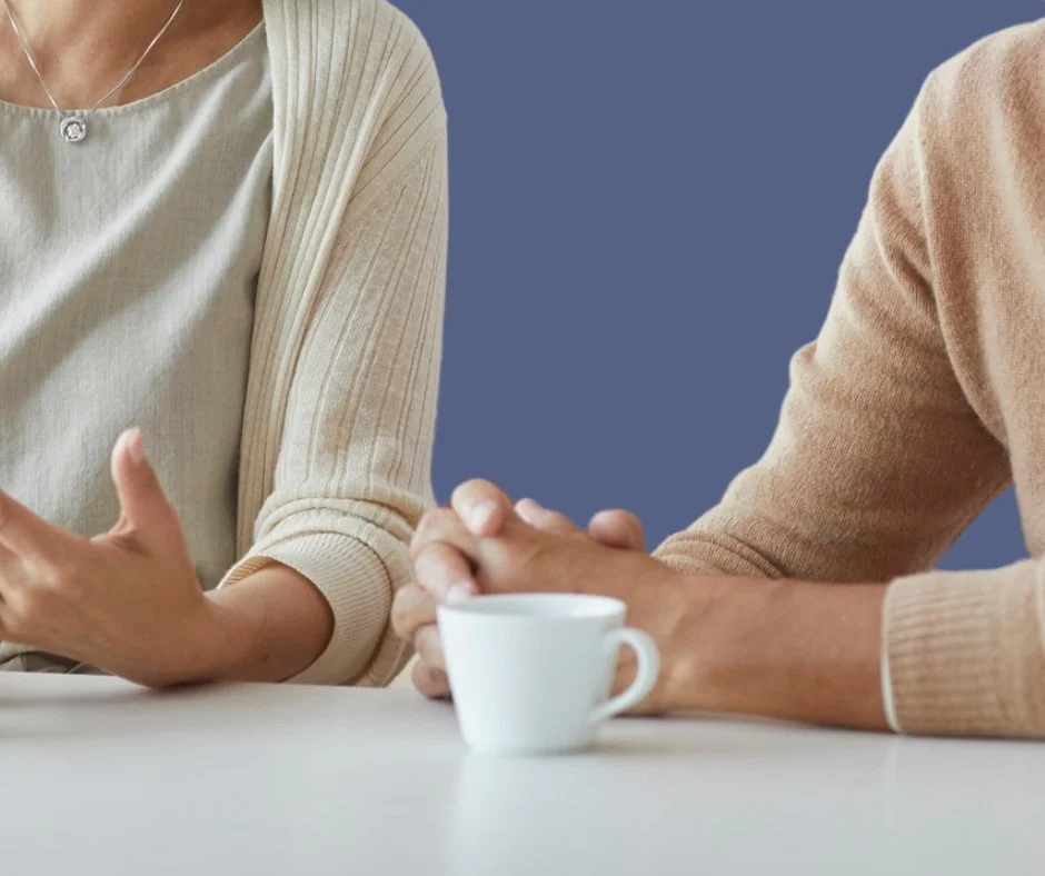 face 2 face counselling. Two people sit at a table, one with their hands gesturing while speaking, and the other with hands clasped near a white coffee cup.