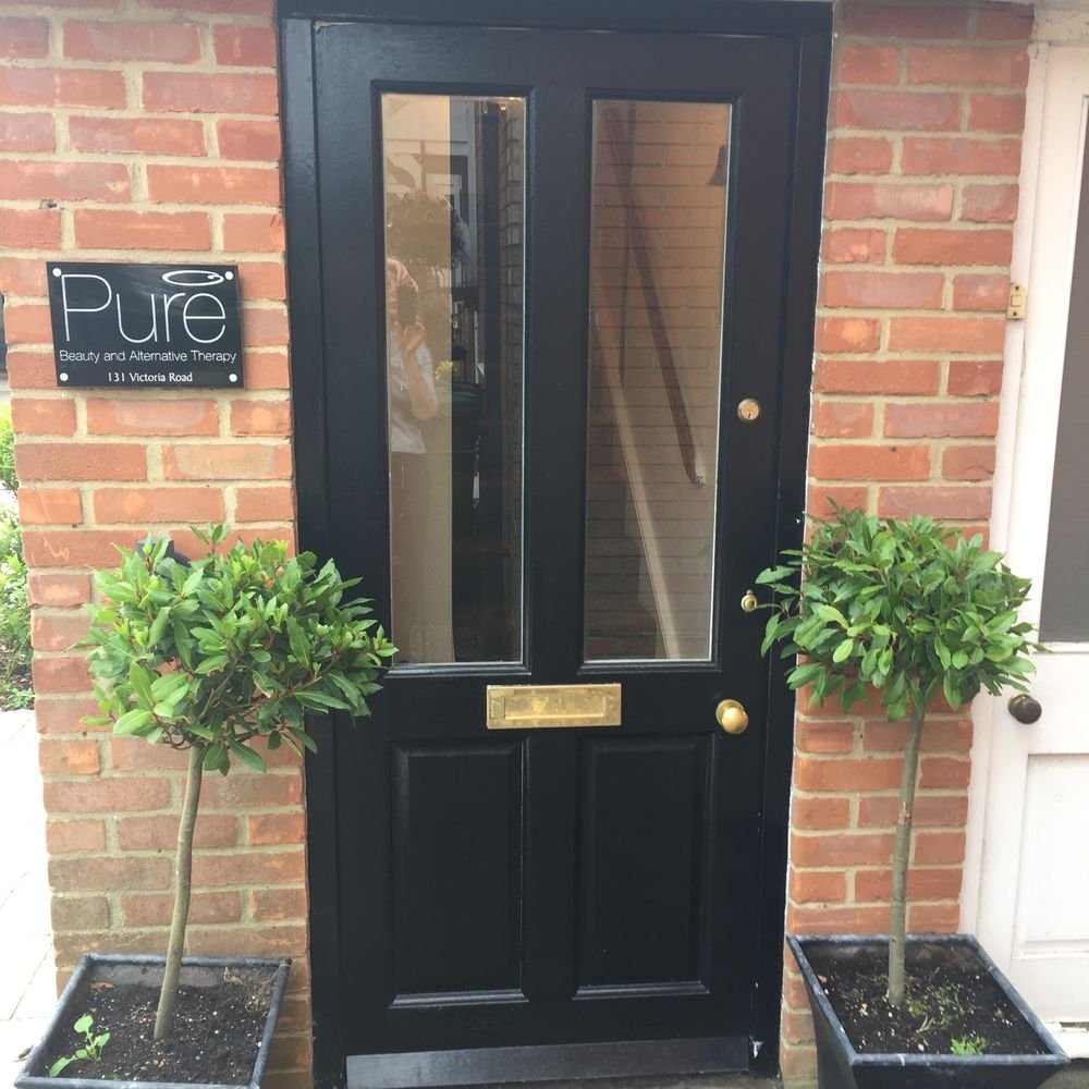 Black front door with glass panels, gold mail slot, and door knob, flanked by two potted topiary trees in square black planters, located on a brick wall exterior.