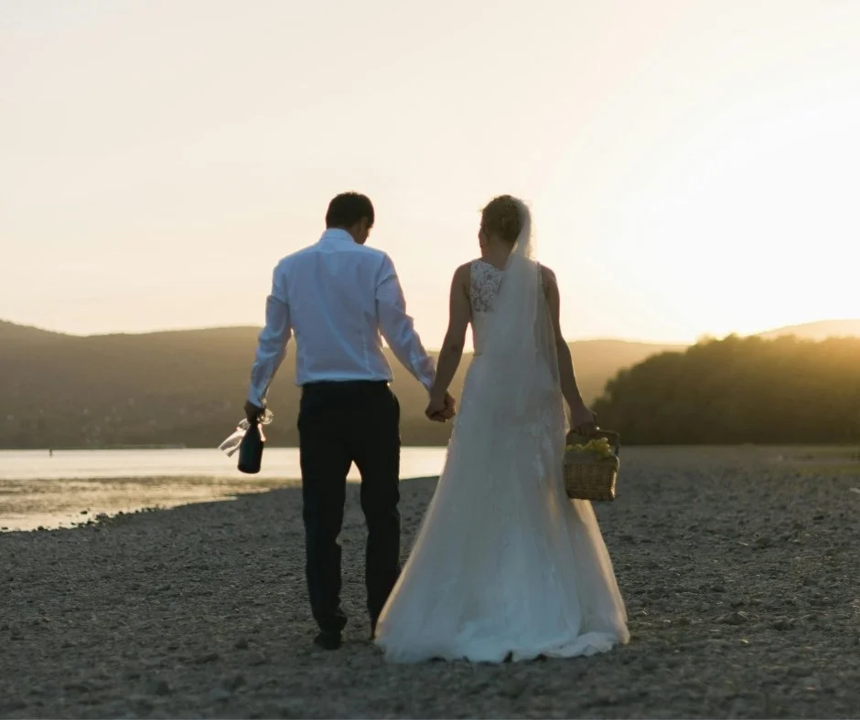 A couple, a bride in a white wedding dress and a groom in a white shirt and dark pants, holding hands and walking on a beach at sunset, with the groom carrying a bottle and a basket, and the bride holding a basket filled with flowers.